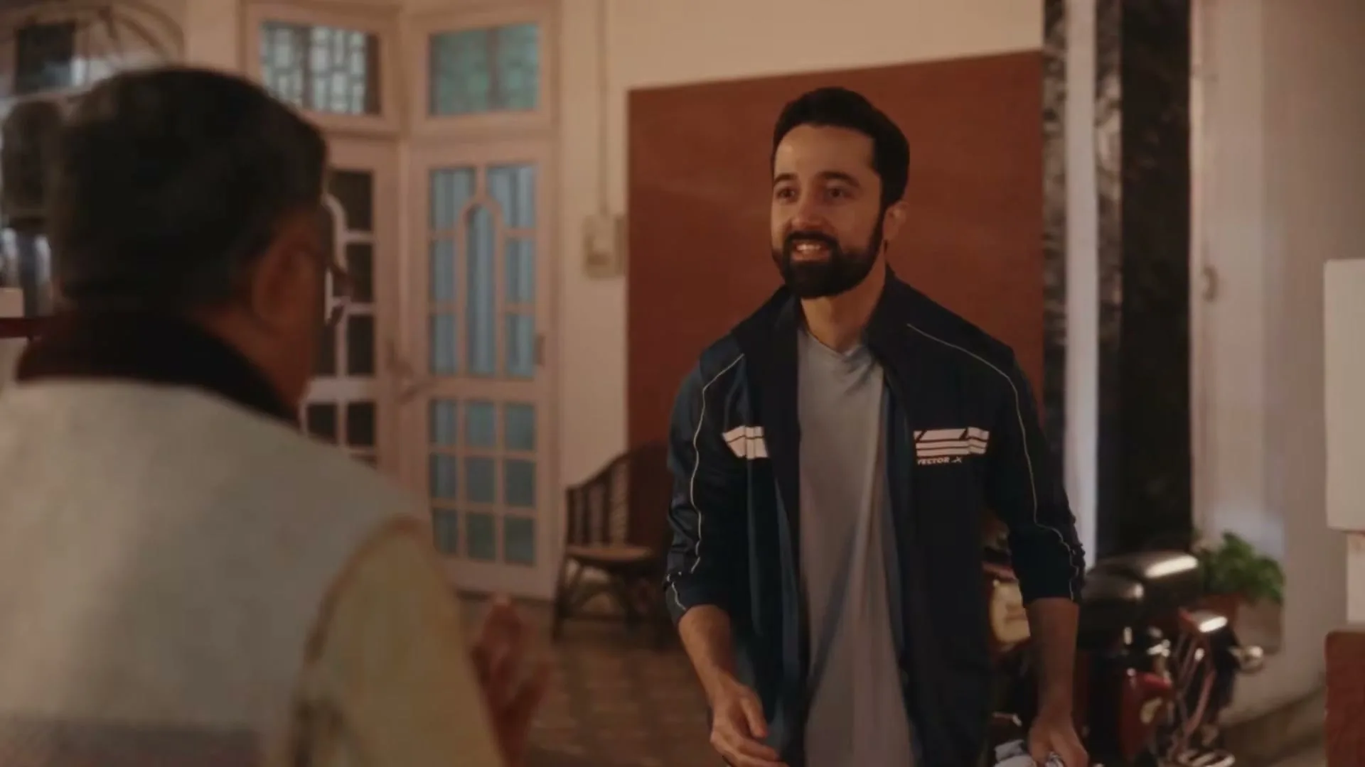 A young man with a beard, wearing a track jacket, smiles at an older man in a home's entryway.