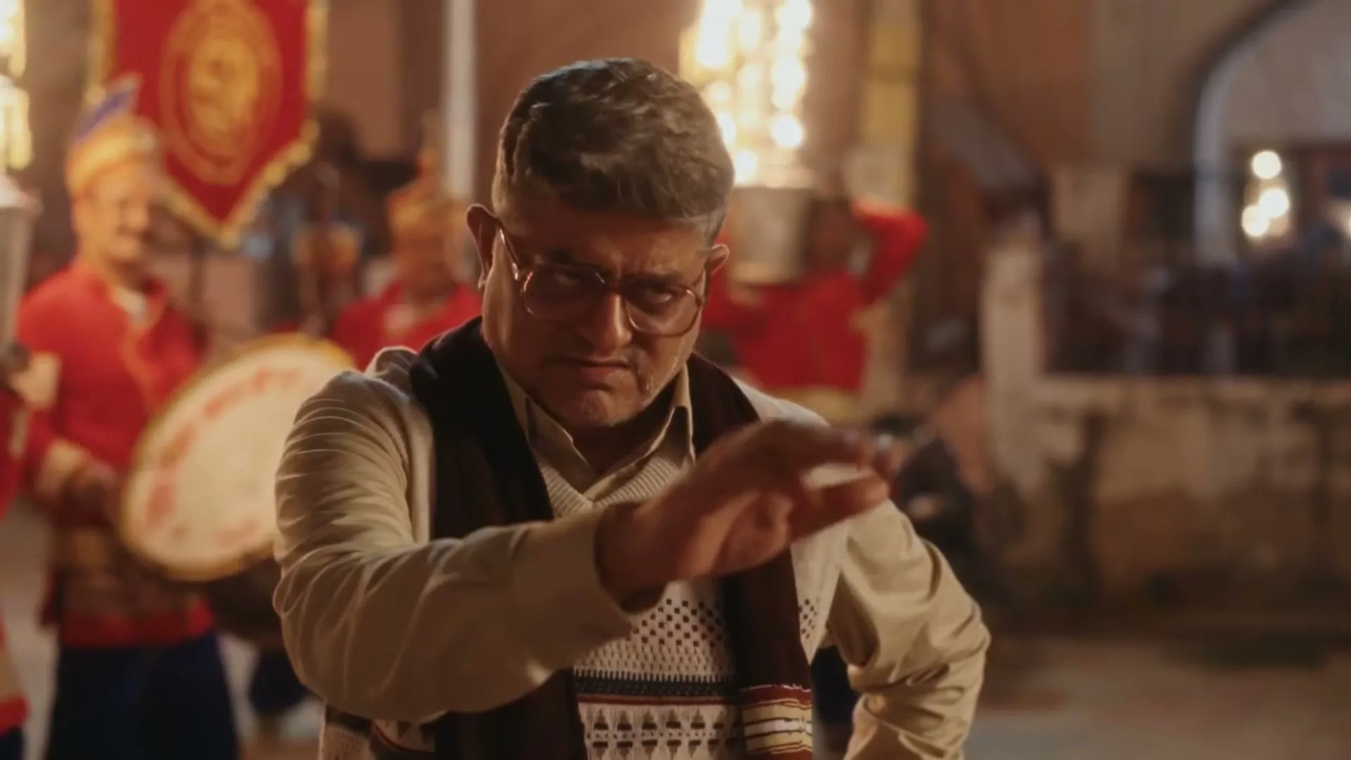 A middle-aged man with glasses and a sweater looks concerned, then gestures with his hand, in a lively outdoor setting with musicians in the background.