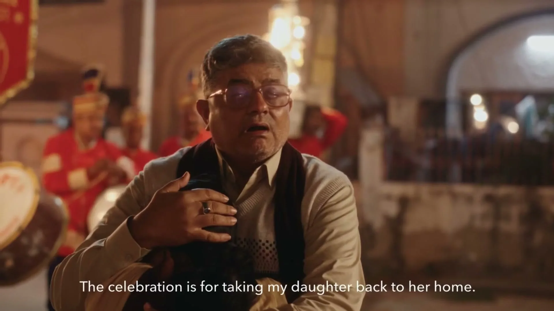 A middle-aged man with glasses and a sweater holds a child close, looking emotional, with a band playing in the background at night.