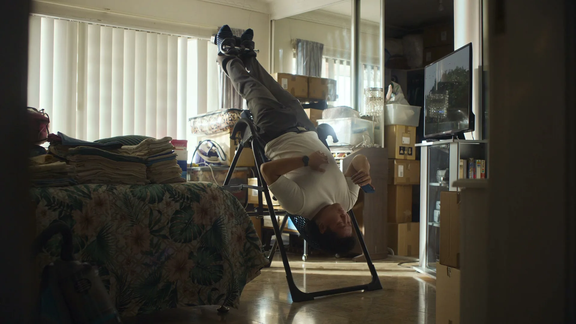 A man in his 30s hangs upside down on an inversion table in a messy bedroom, looking at his phone.