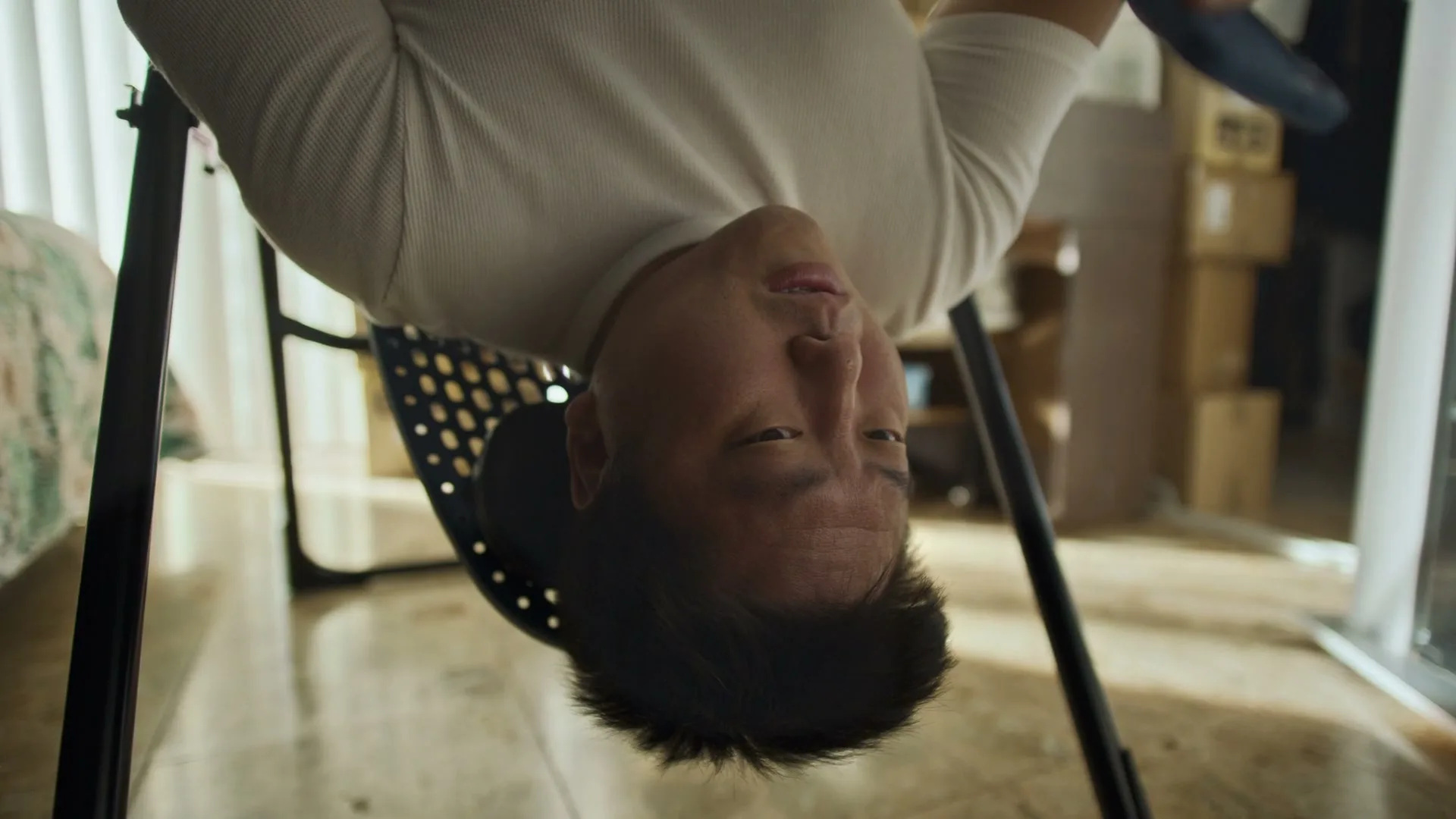 A man hangs upside down from an inversion table, looking at the camera with a slightly concerned expression in a brightly lit room.
