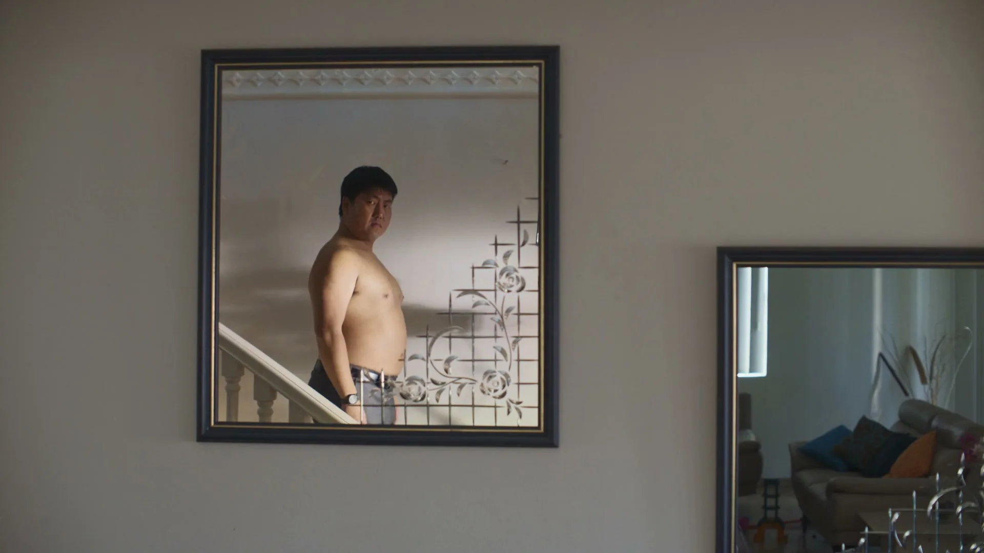 A shirtless man in his 30s stands in front of a mirror, looking at his reflection in a home interior.