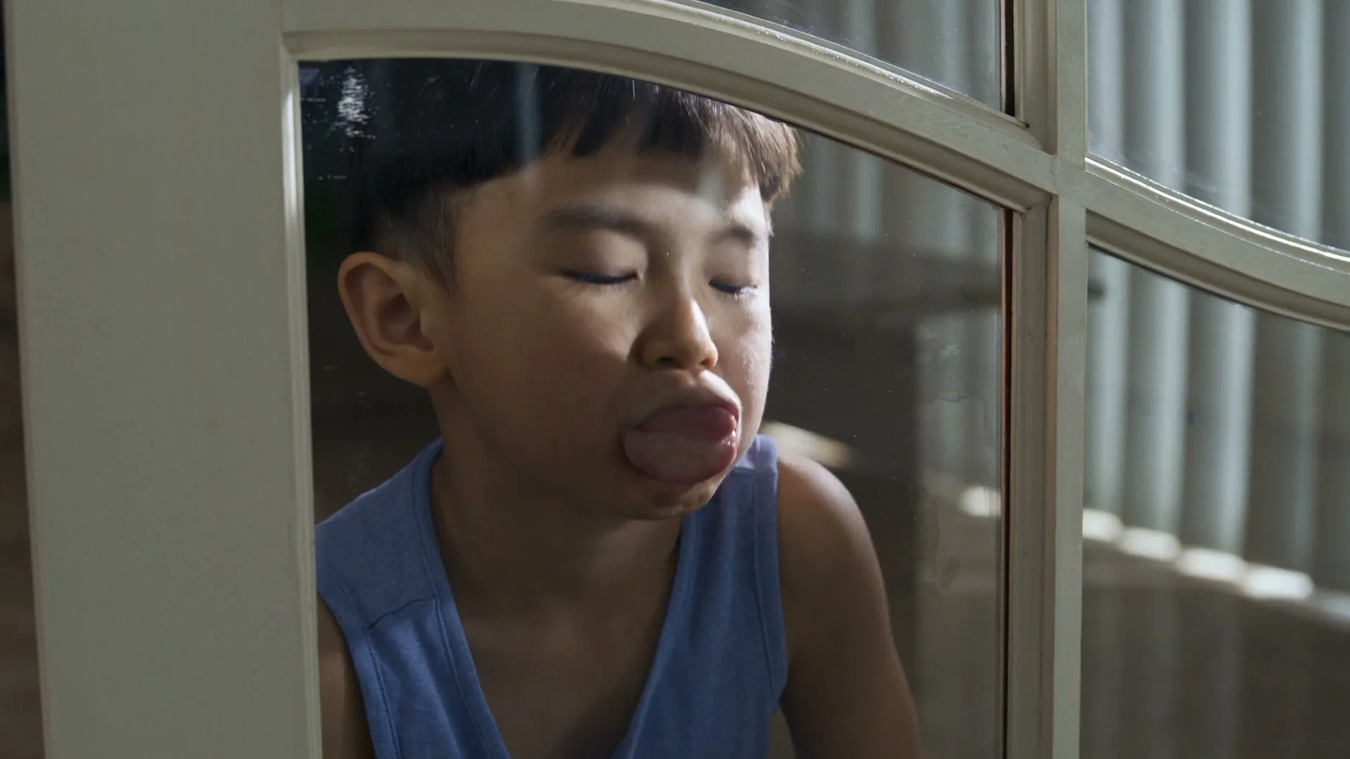 A young boy with dark hair and a blue tank top presses his face against a glass window, making silly expressions.