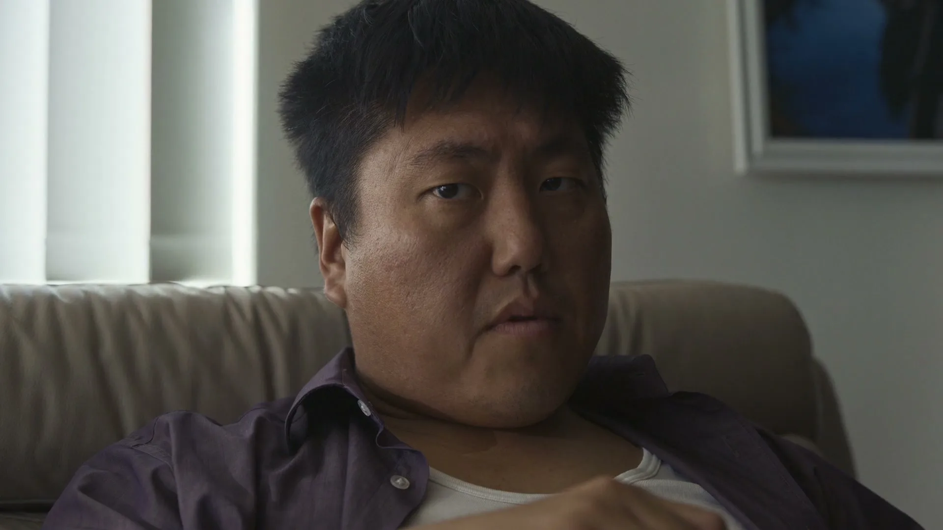 A middle-aged Asian man with dark hair and a purple shirt sits on a couch, looking directly at the camera with a serious expression in an interior setting.