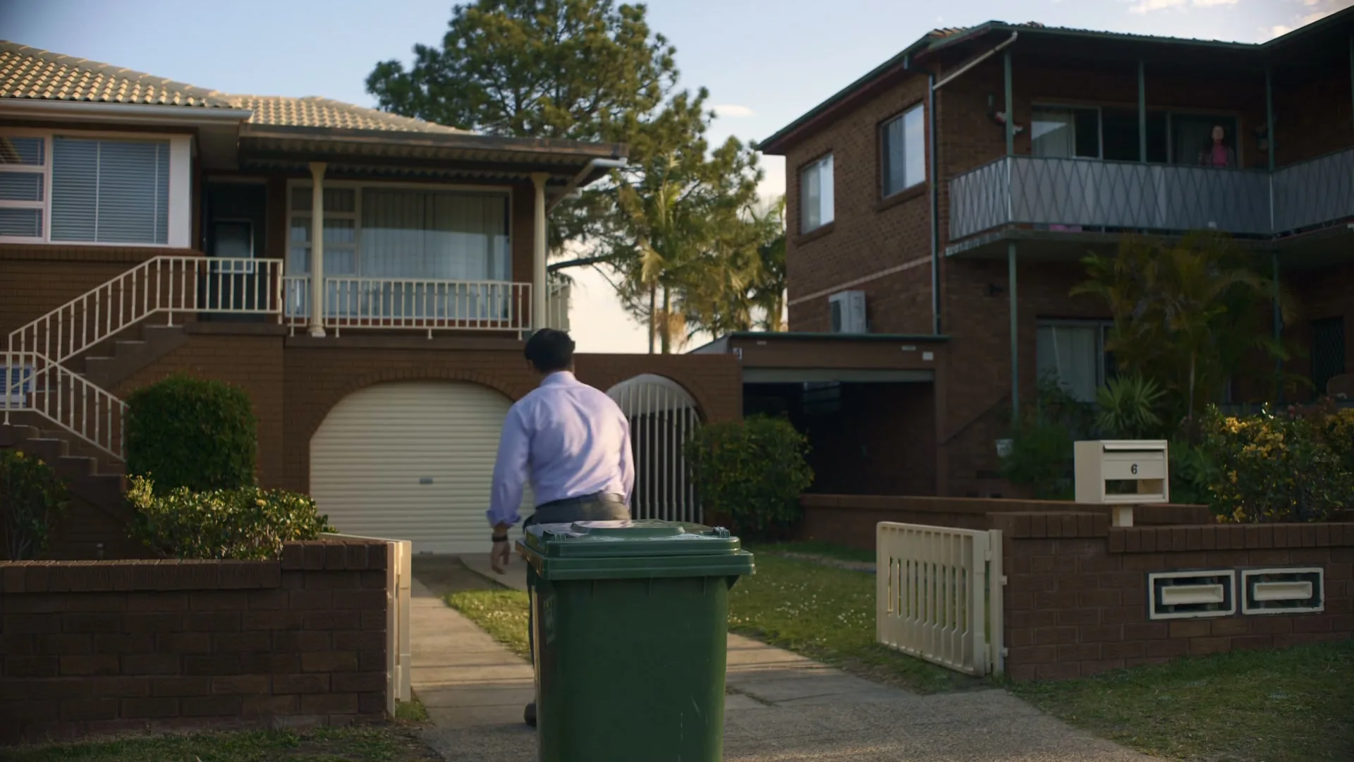 A man in a shirt and tie pushes a green wheelie bin down a driveway in a suburban street.