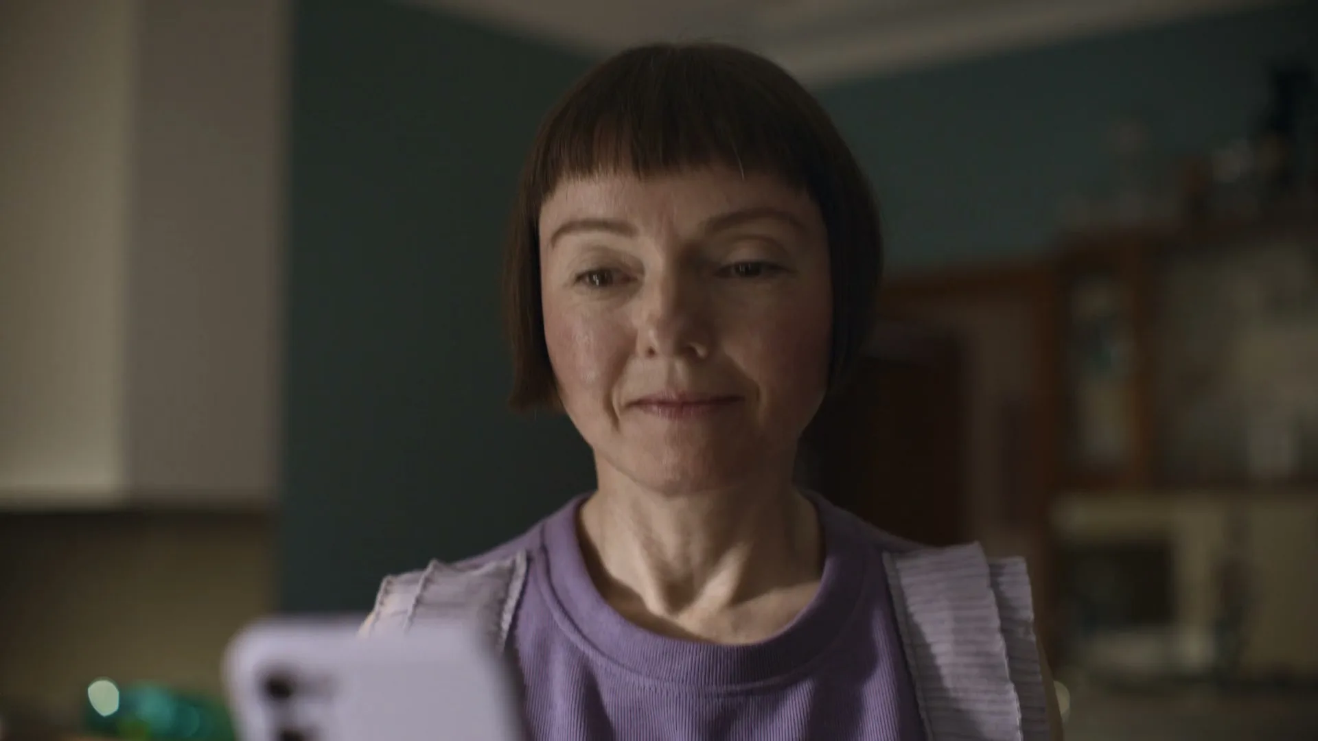 A woman with a bob haircut and bangs, wearing a purple top, looks down at her phone in a dimly lit kitchen.