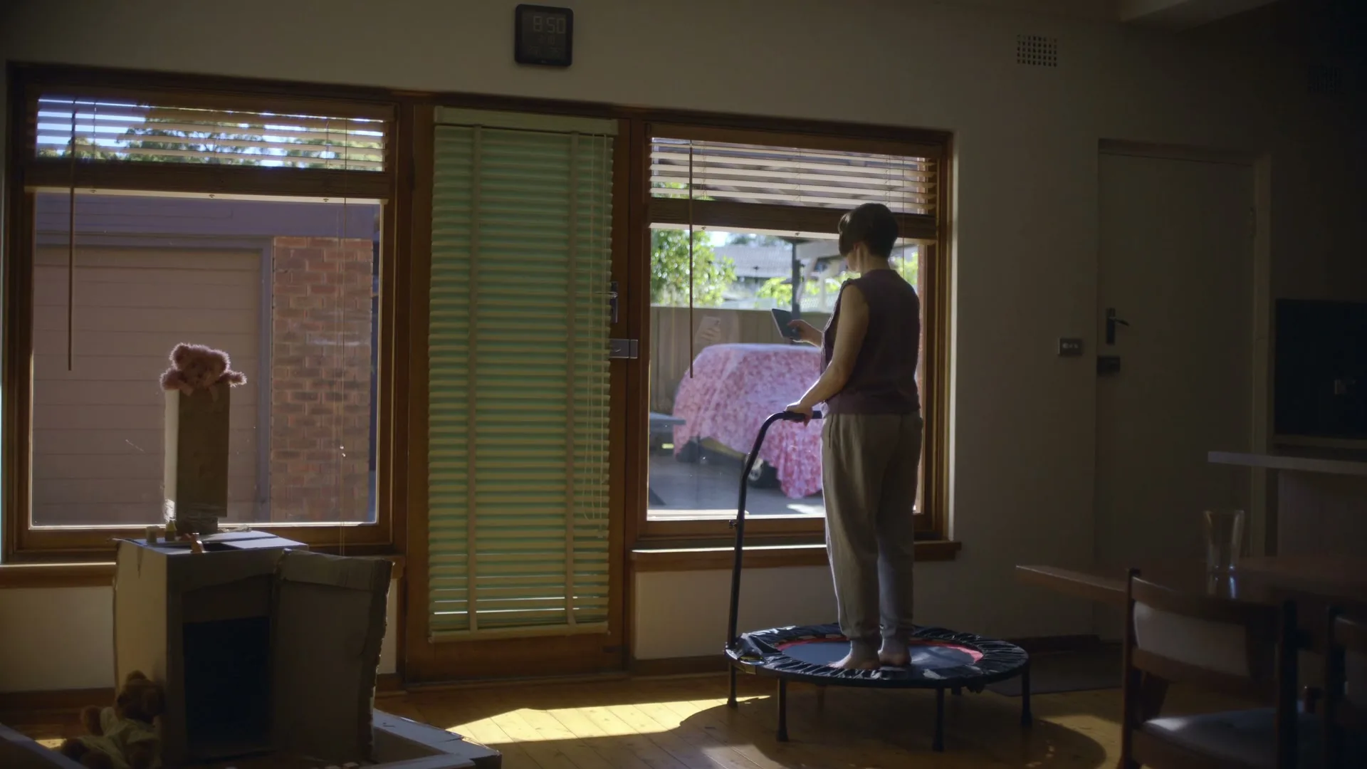 A woman in casual clothes stands on a mini trampoline, looking out a window in a sunlit living room.