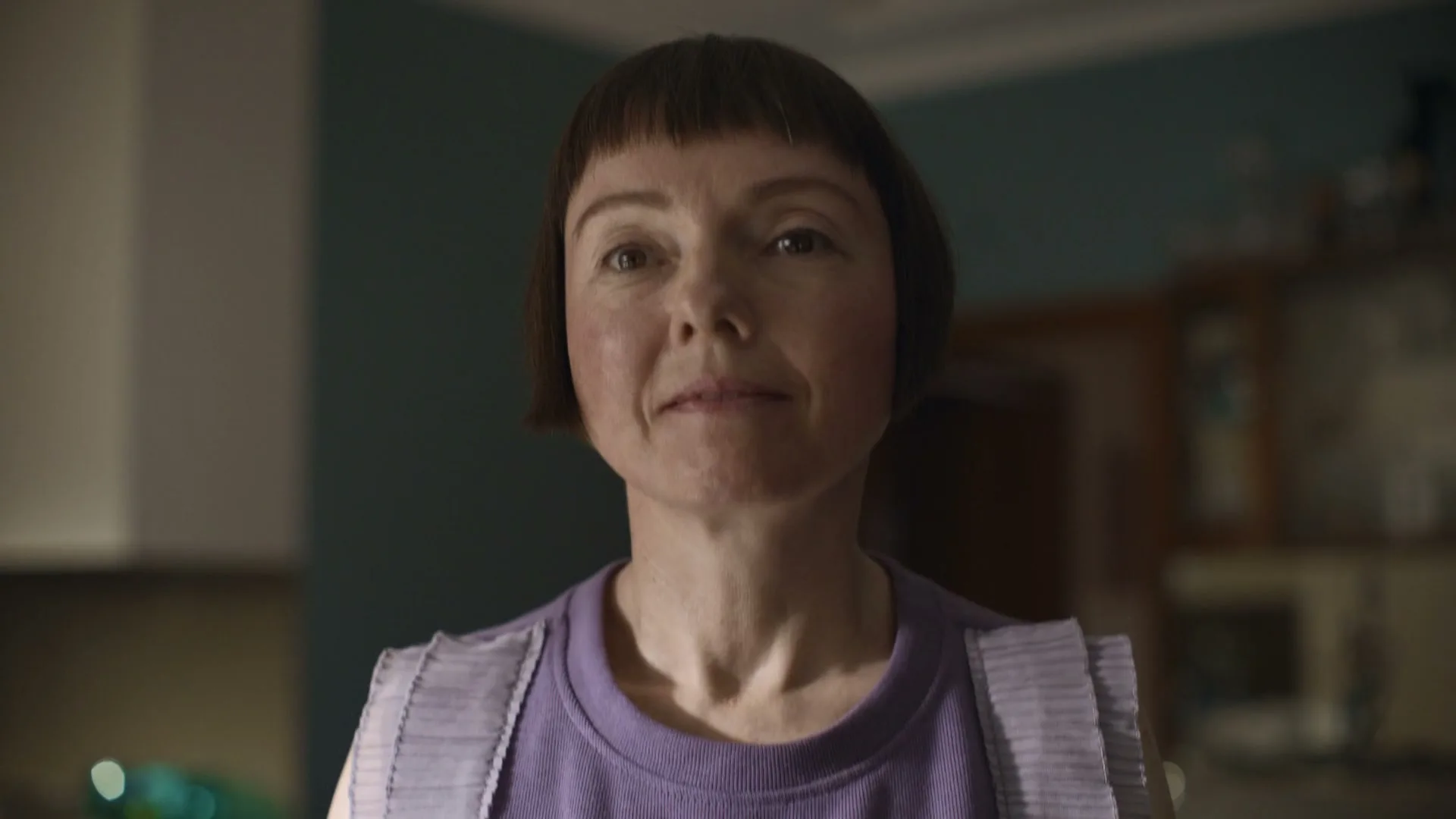 A woman with a bob haircut and purple shirt looks up with a slight smile in a dimly lit kitchen.