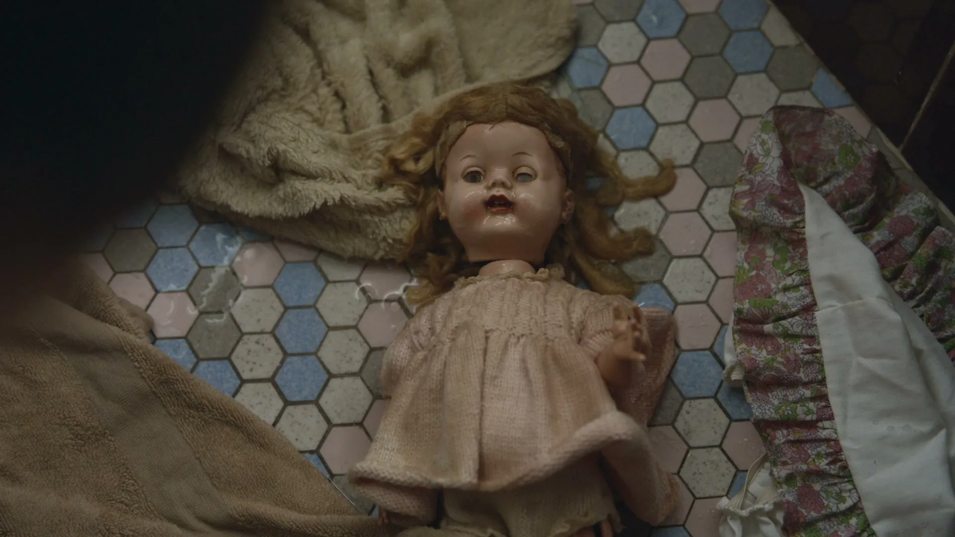 A person's hand pulls away a dark blue cloth to reveal an old, creepy doll lying on a tiled floor.