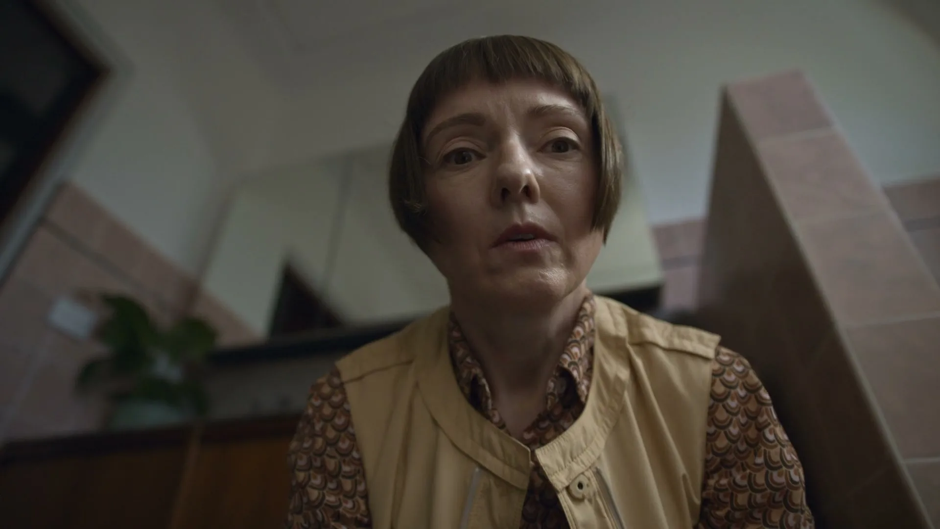 A woman with a bob haircut and a patterned shirt looks directly at the camera with a concerned expression in a tiled room.