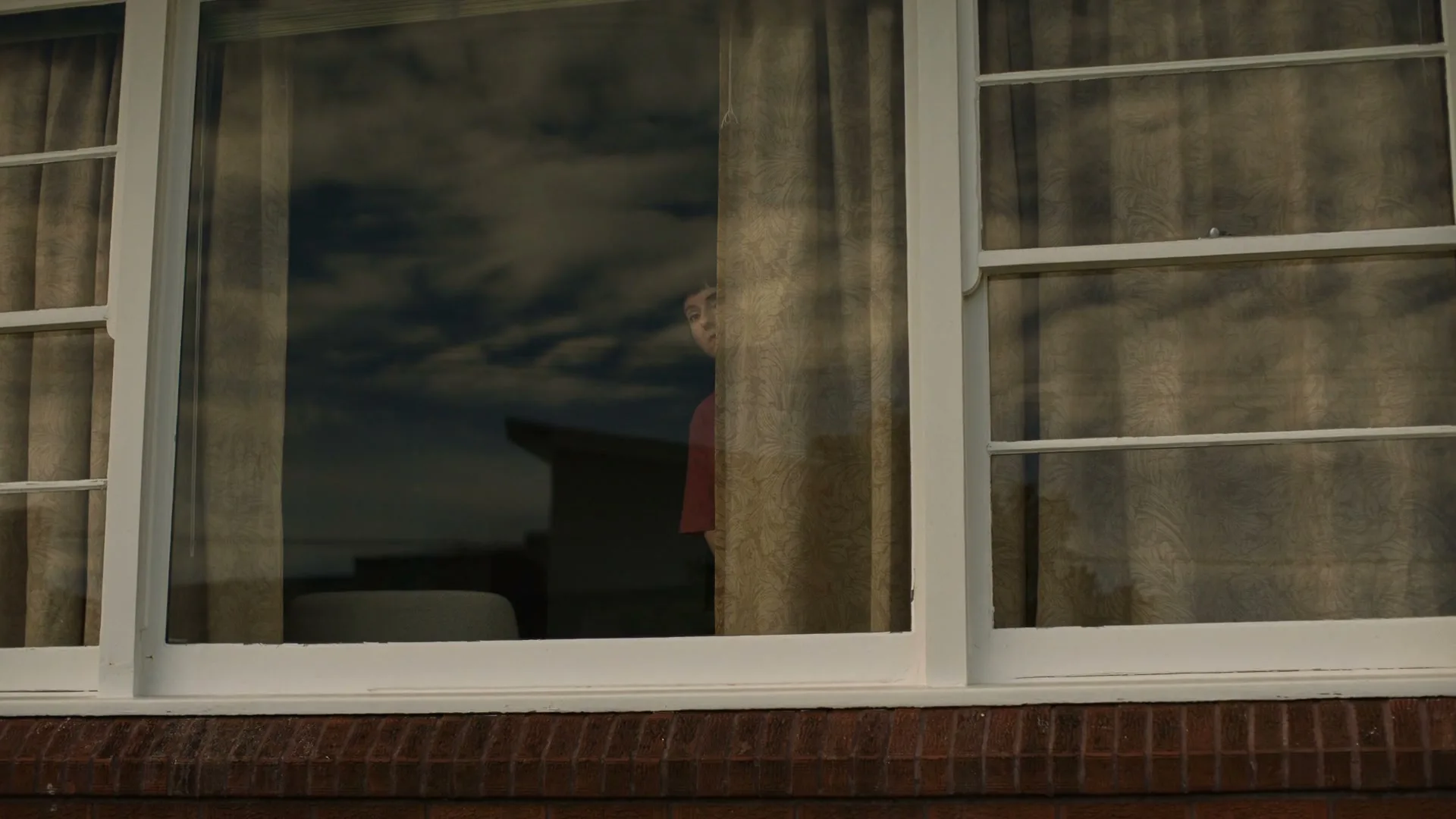 A young woman with dark hair and a red shirt peeks out from behind a curtain in a window, looking surprised, in a suburban house during the day.