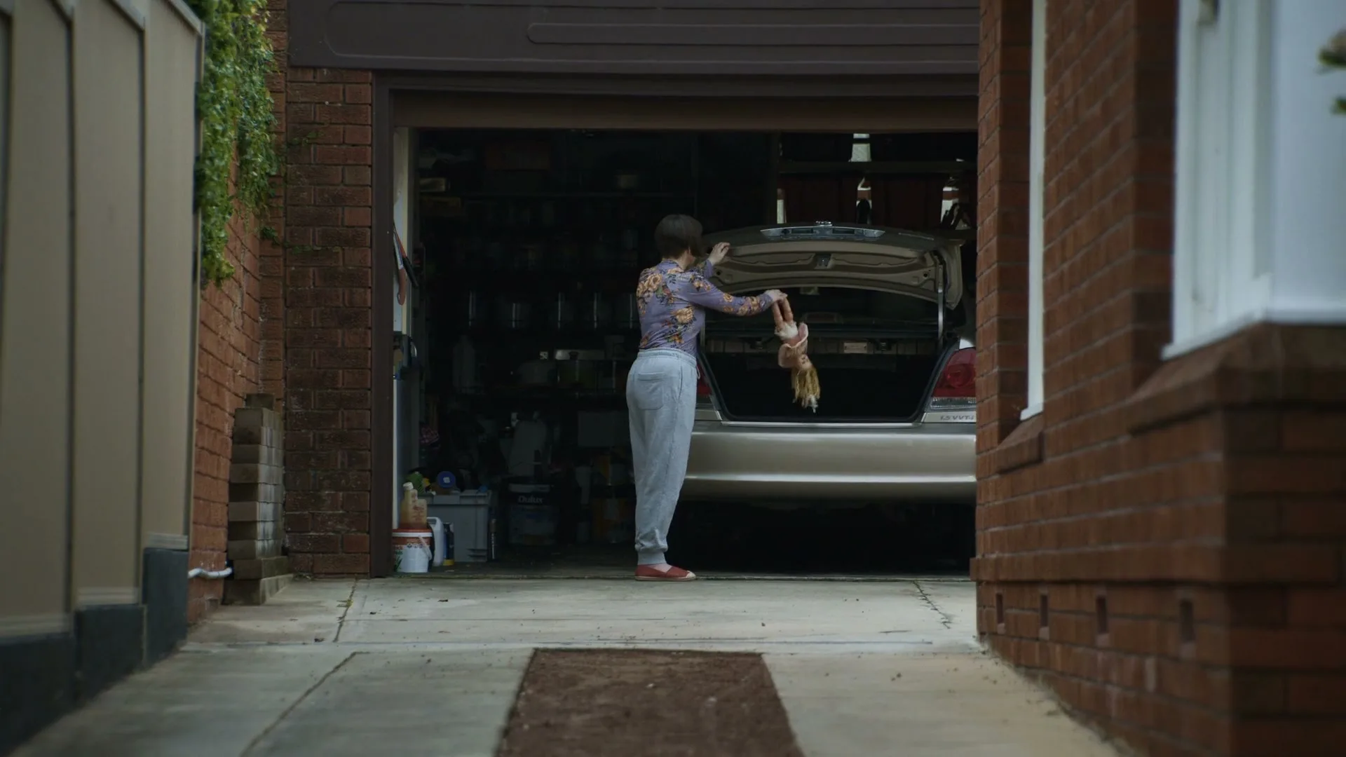 A woman with short brown hair, wearing a patterned top and grey pants, stands in a garage driveway, putting a doll into the trunk of a car.