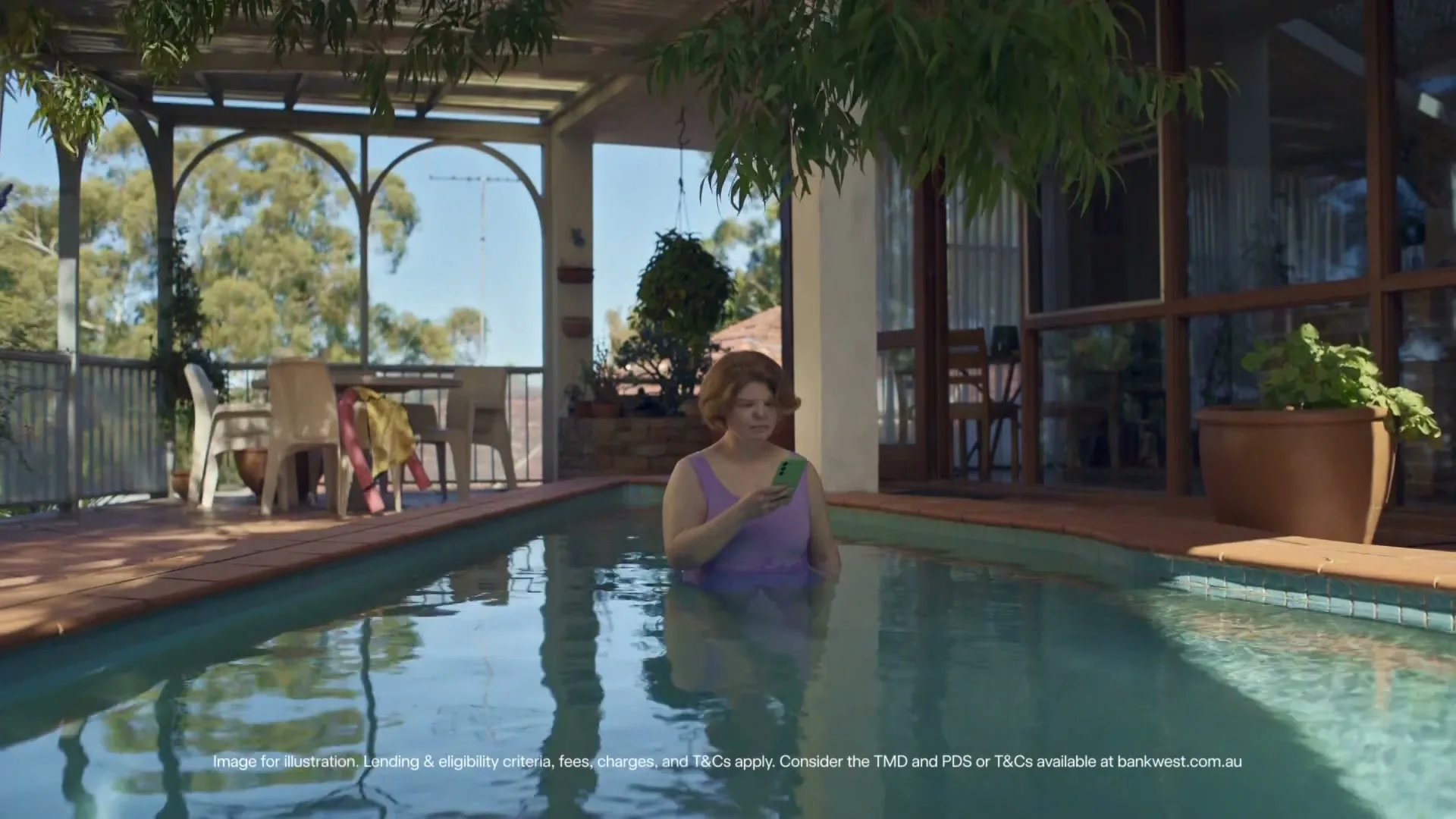 A woman in a purple swimsuit stands in a swimming pool, looking at her phone on a sunny day in a suburban backyard.