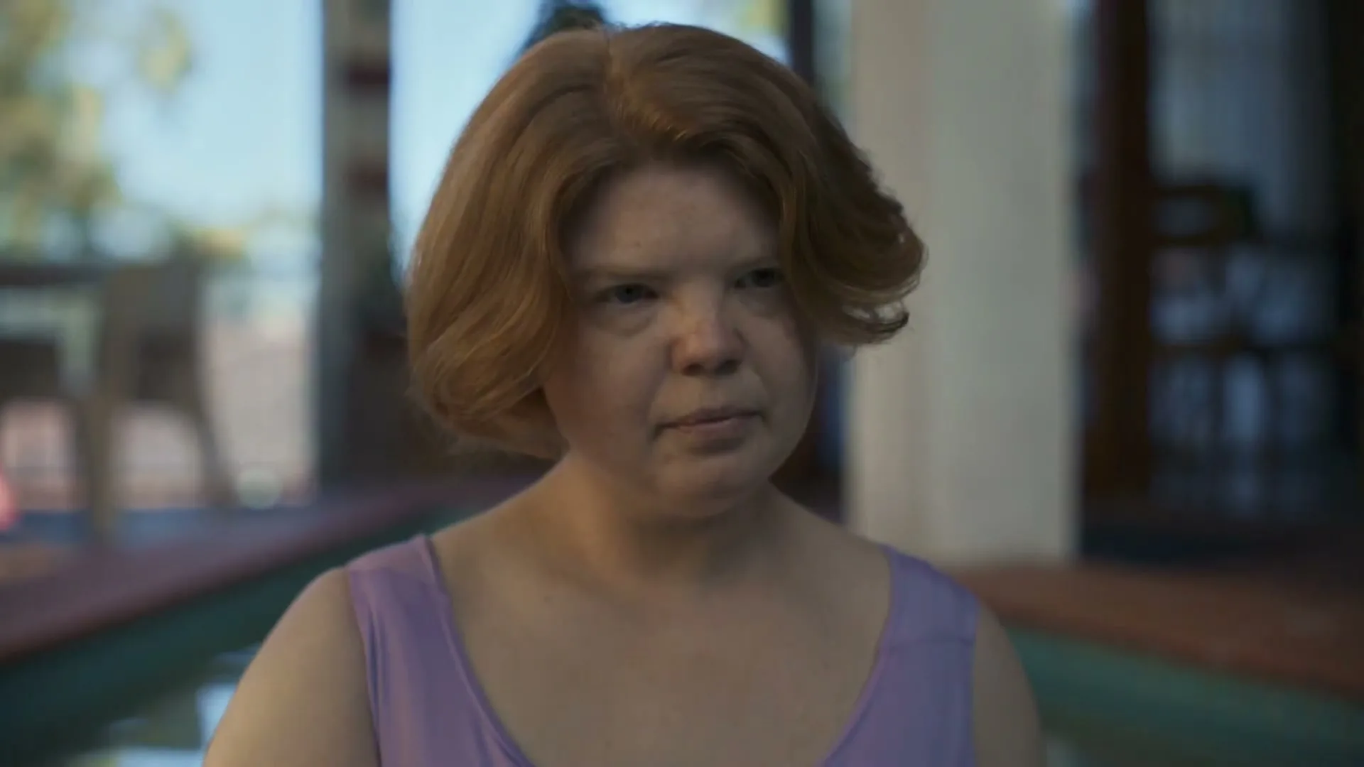 A woman with red hair and a purple top looks at her phone, then up at the camera, in a bright, modern interior with a pool.