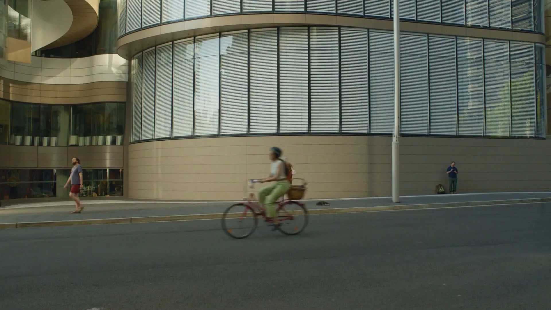 A woman on a bicycle rides past a modern building, while a man walks and another man stands on the sidewalk.