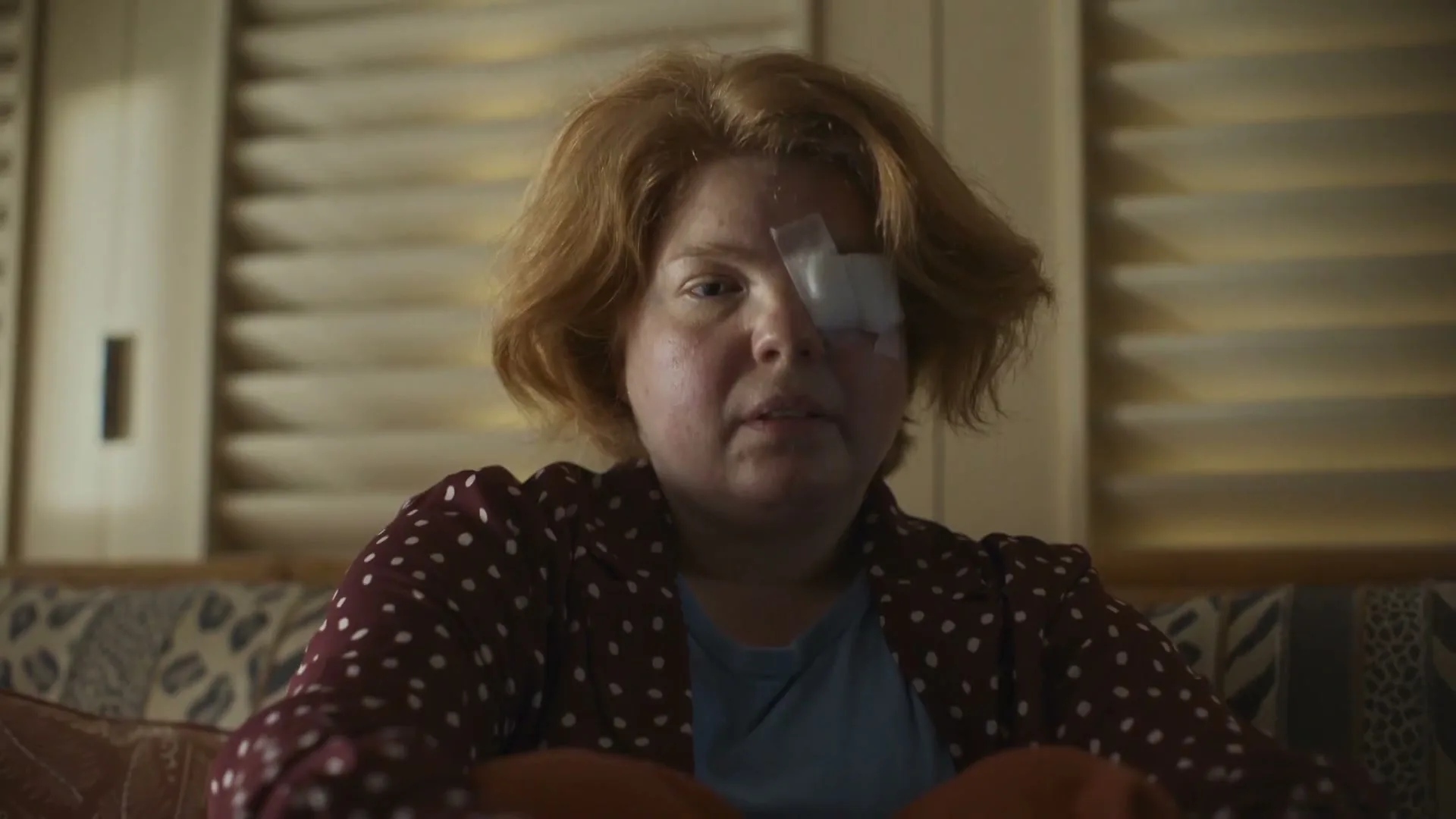 A woman with red hair and an eye patch sits on a patterned couch in a dimly lit room, looking directly at the camera.