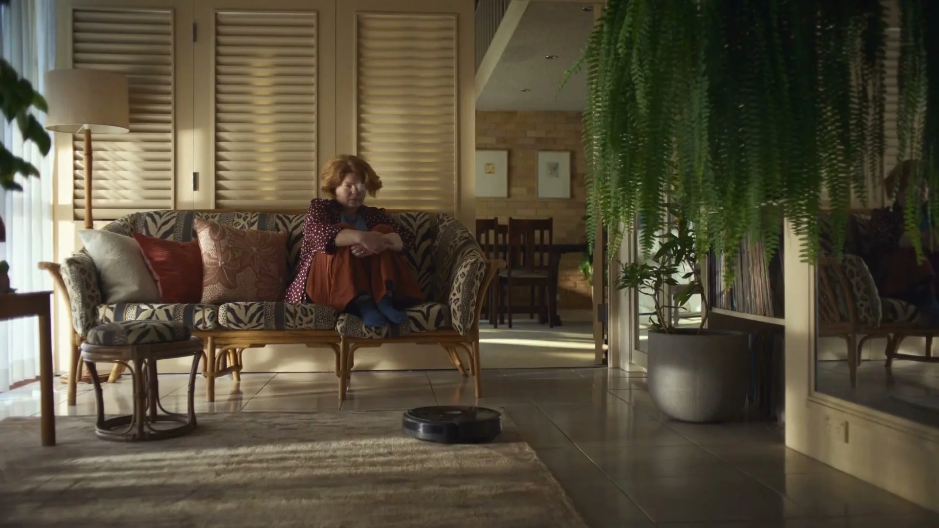 A woman with red hair sits on a patterned couch, looking down at a robot vacuum cleaner moving on the rug in a sunlit living room.