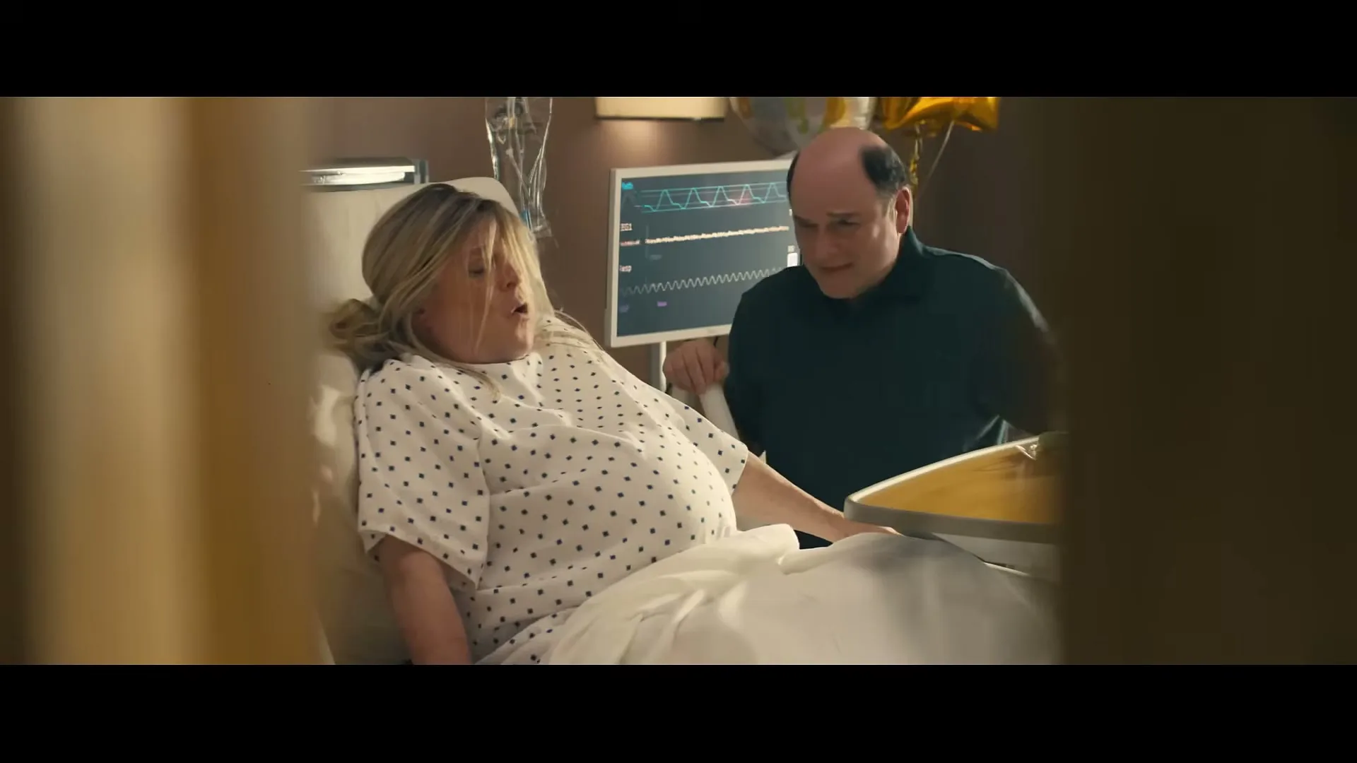 A pregnant woman in a hospital gown is in labor, pushing in a hospital bed, while a bald man in a dark polo shirt stands beside her, looking concerned.
