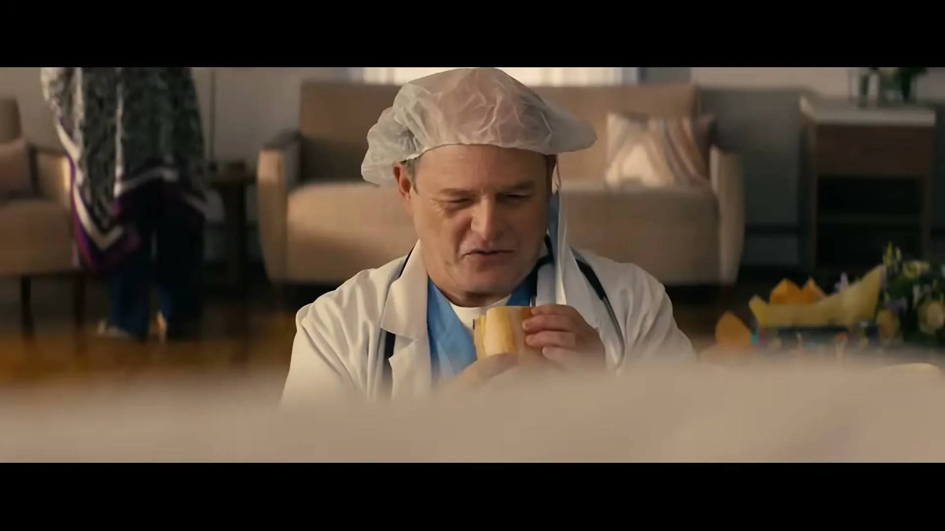 A male doctor in scrubs and a hairnet sits on the floor, eating a sandwich in a living room.