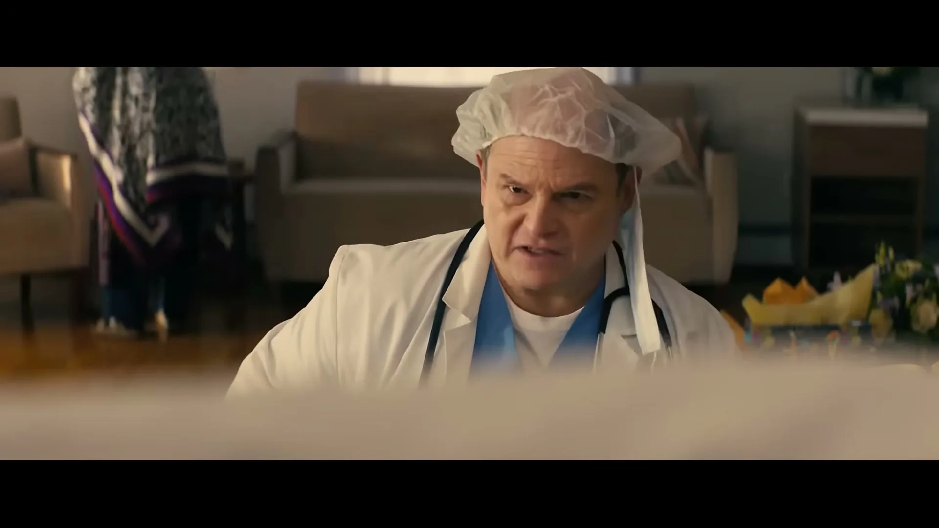 A male doctor in a surgical cap and white coat looks down at a baby, then smiles and holds up a bottle of Mike's Amazing mayonnaise in a living room.