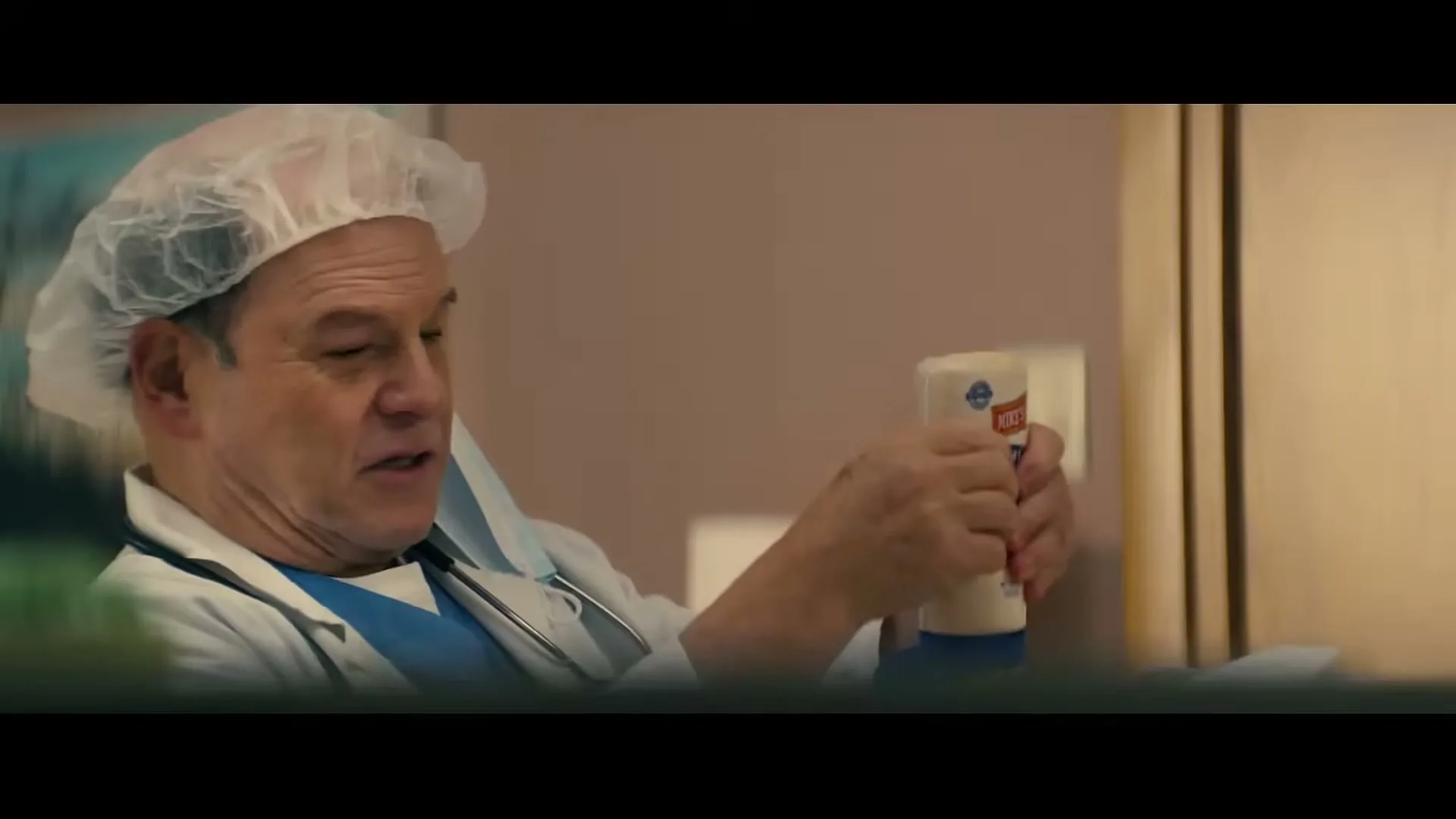 A male doctor in scrubs and a hairnet holds a bottle of Mike's Amazing mayonnaise, looking displeased in a hospital room.