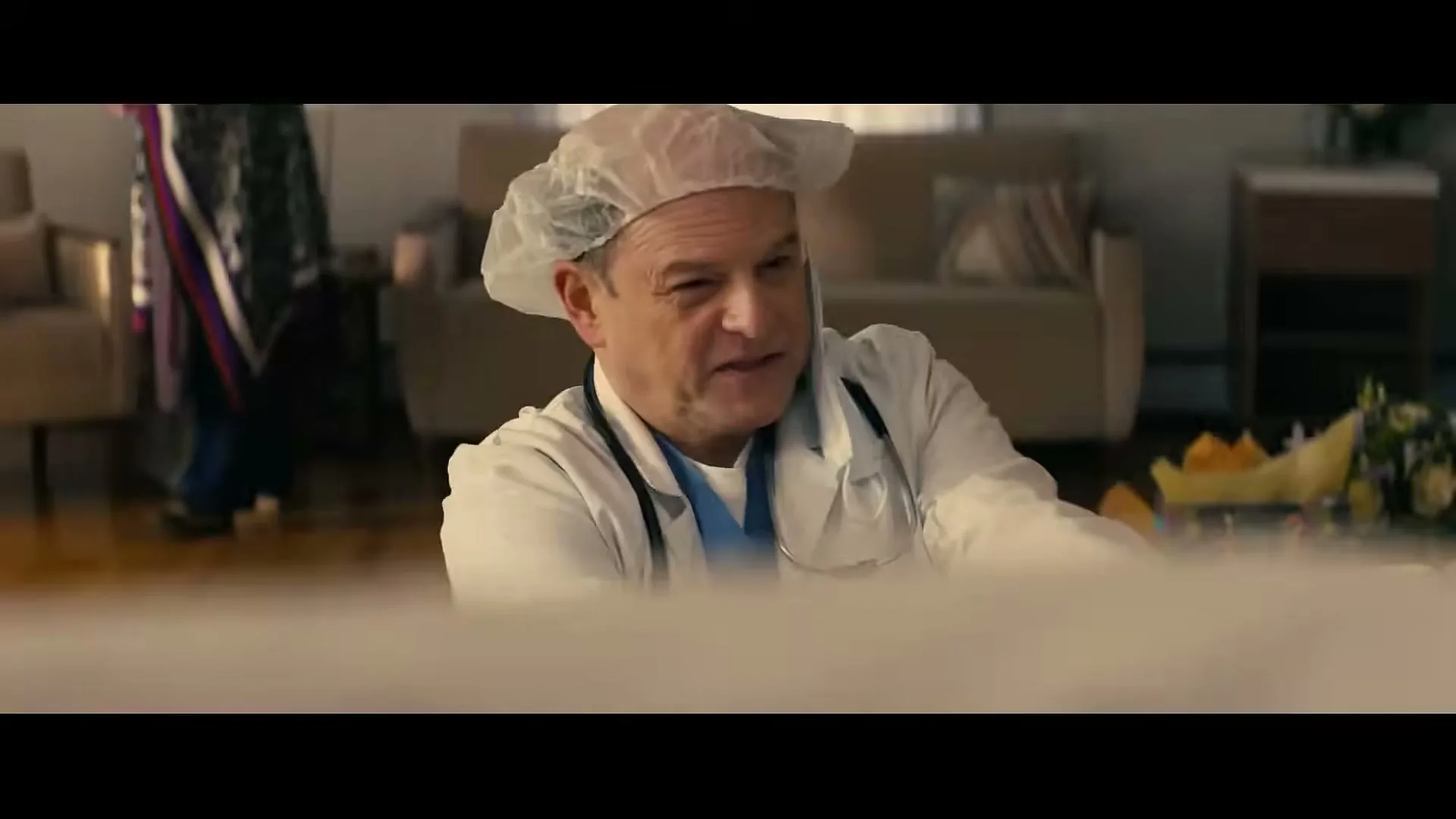 A middle-aged man in a doctor's coat and hairnet looks down and smiles, with a living room in the background.
