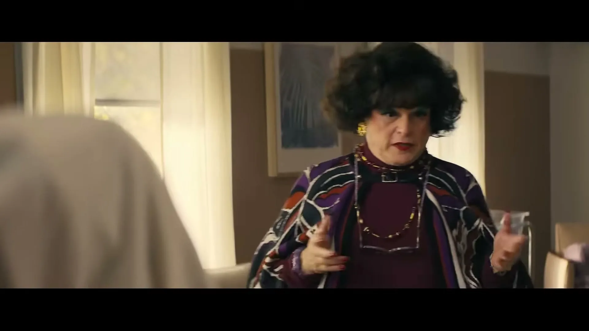 A person in drag, dressed in a purple top and patterned cape, with a dark wig and heavy makeup, gestures while speaking in an interior setting.