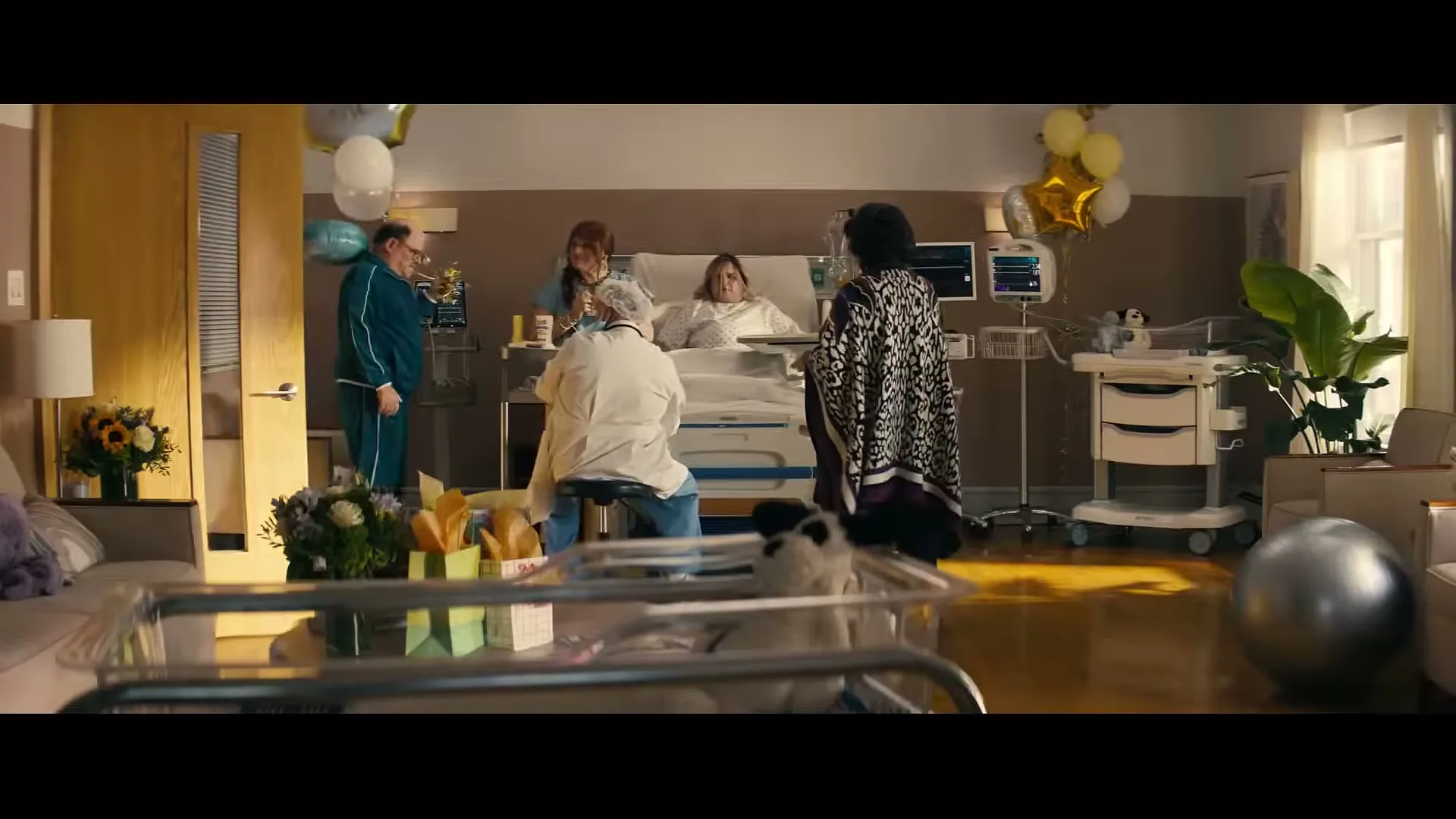 A new mother lies in a hospital bed surrounded by family and friends, including a man in a blue tracksuit and a woman in a patterned coat, in a brightly lit hospital room.