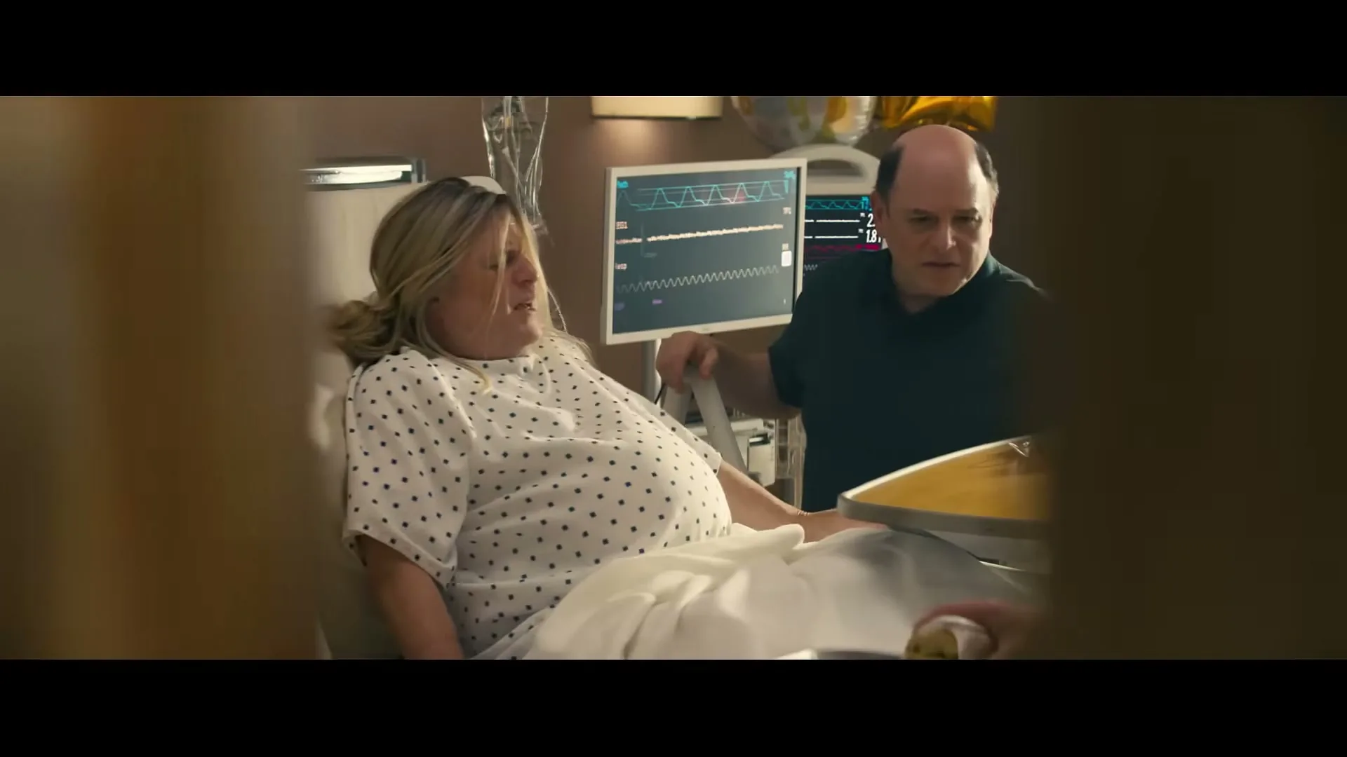 A pregnant woman in a hospital gown lies in a bed while a man in a dark shirt stands beside her, looking at a medical monitor in a hospital room.