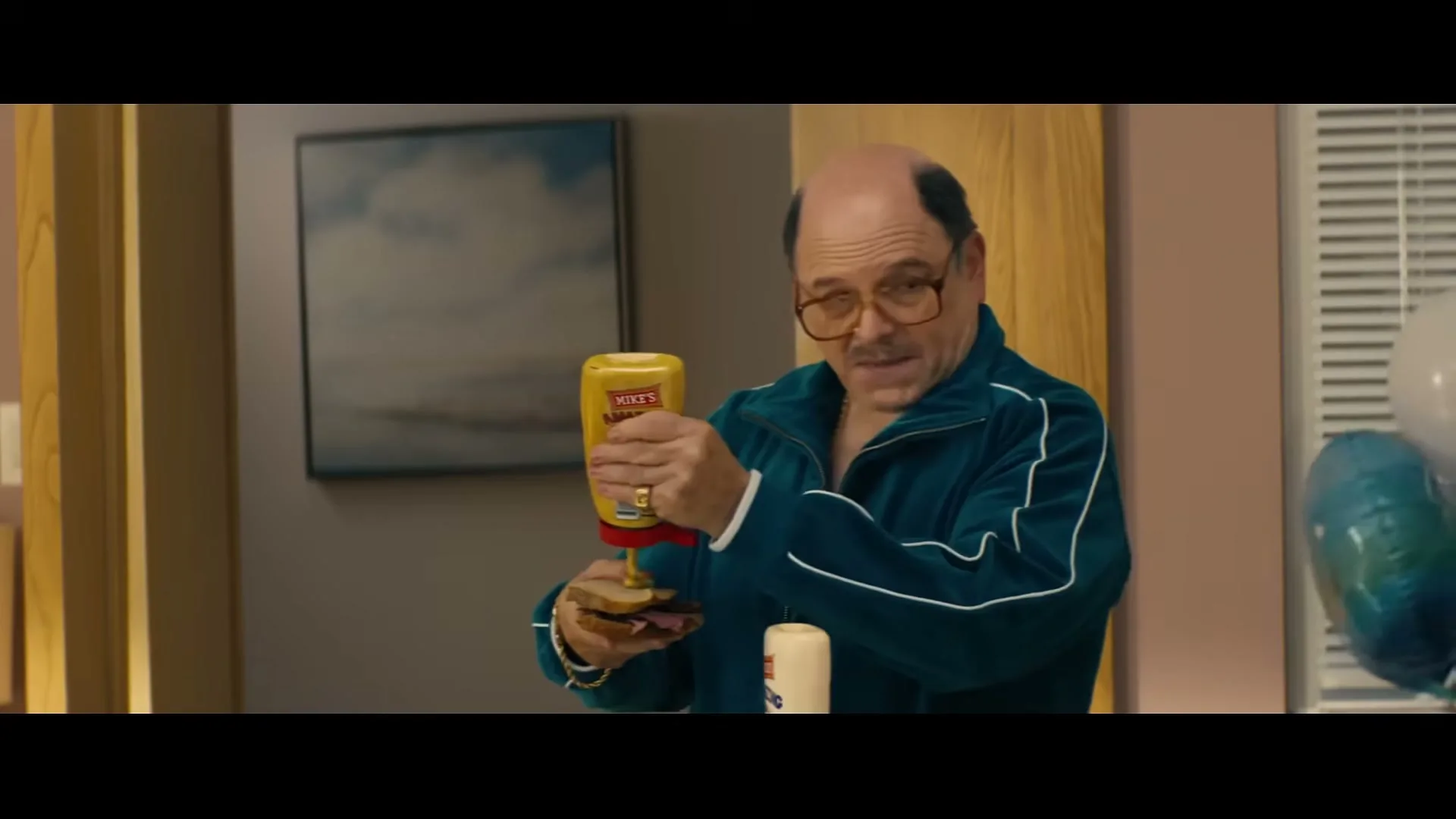 A bald man in a teal tracksuit squeezes mustard onto a sandwich in an interior setting.