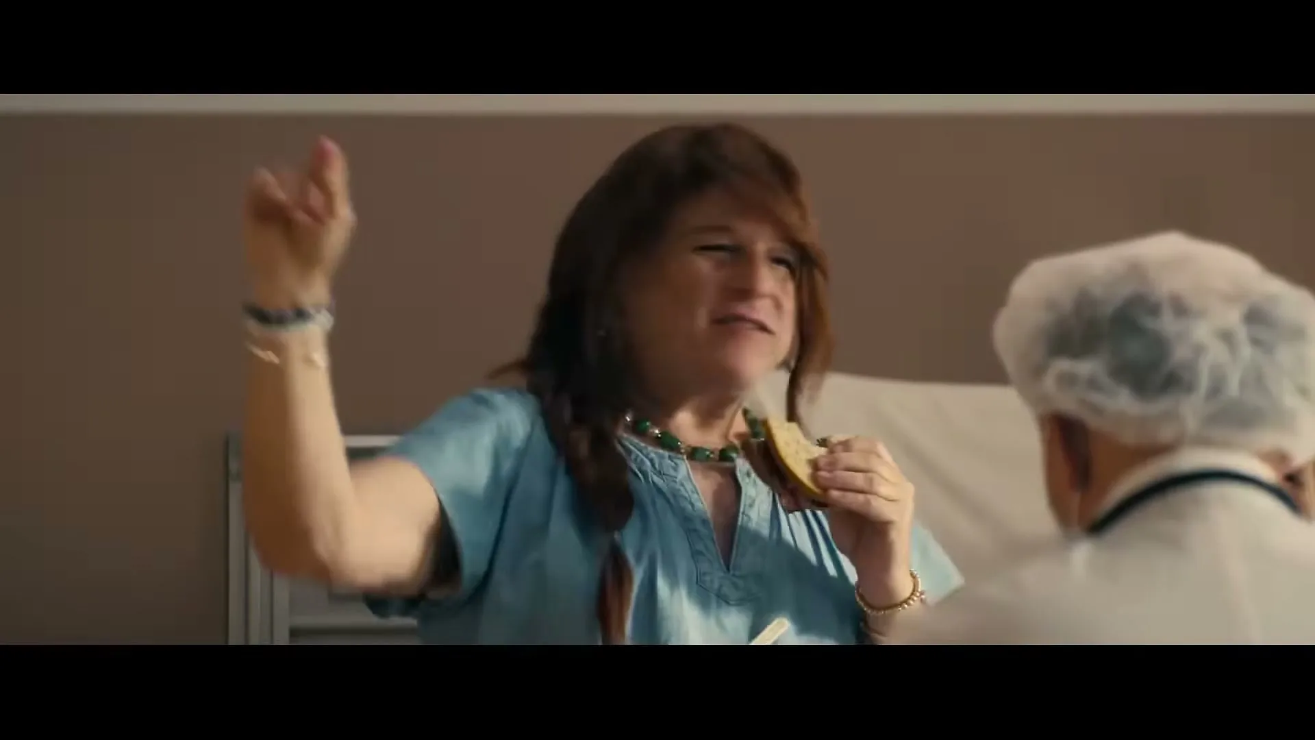 A person in a wig and hospital gown eats a cracker and gestures while talking to a person in a hairnet in a hospital room.