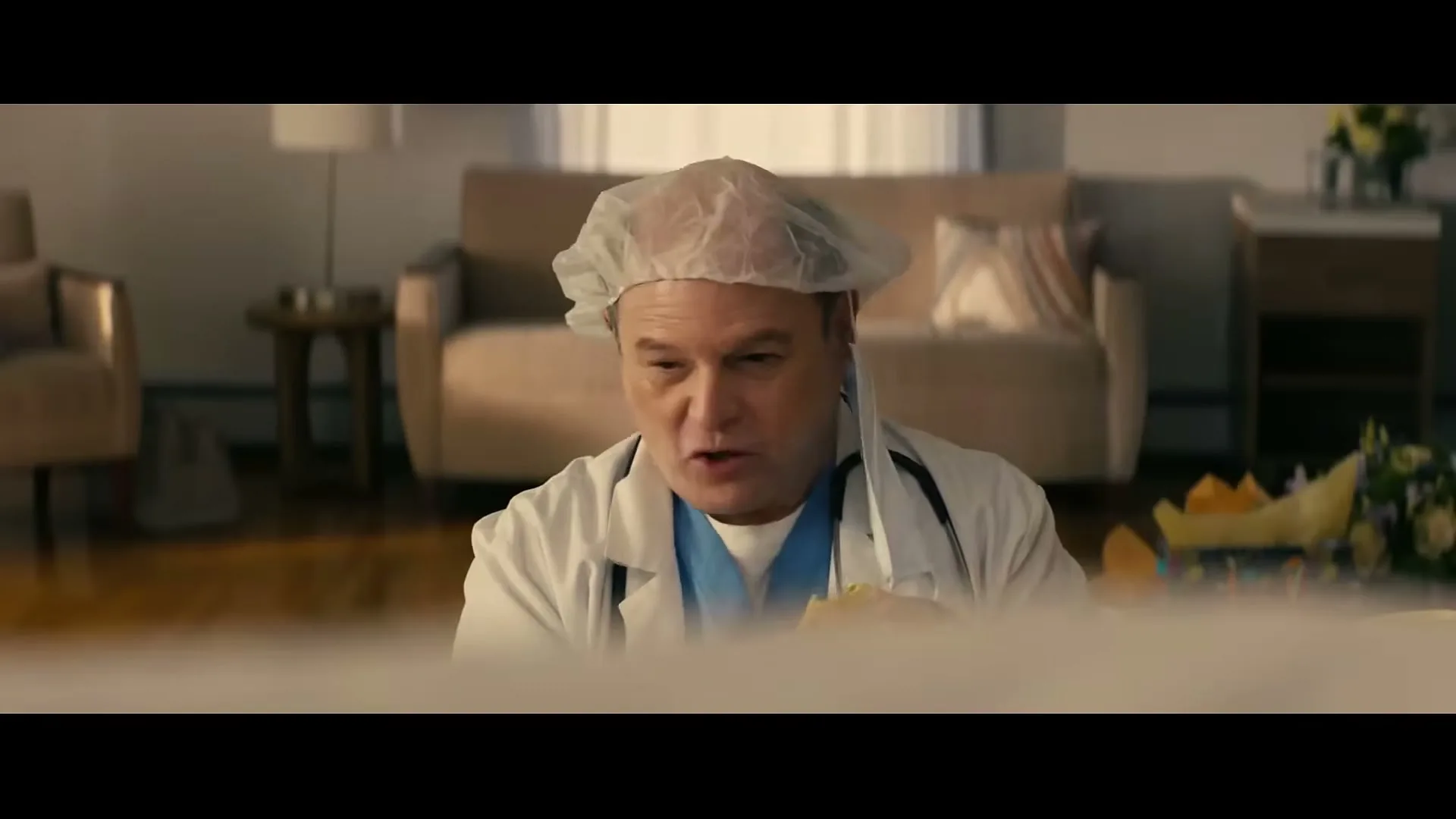 A male doctor in scrubs and a hairnet looks down with a disgusted expression in a living room.