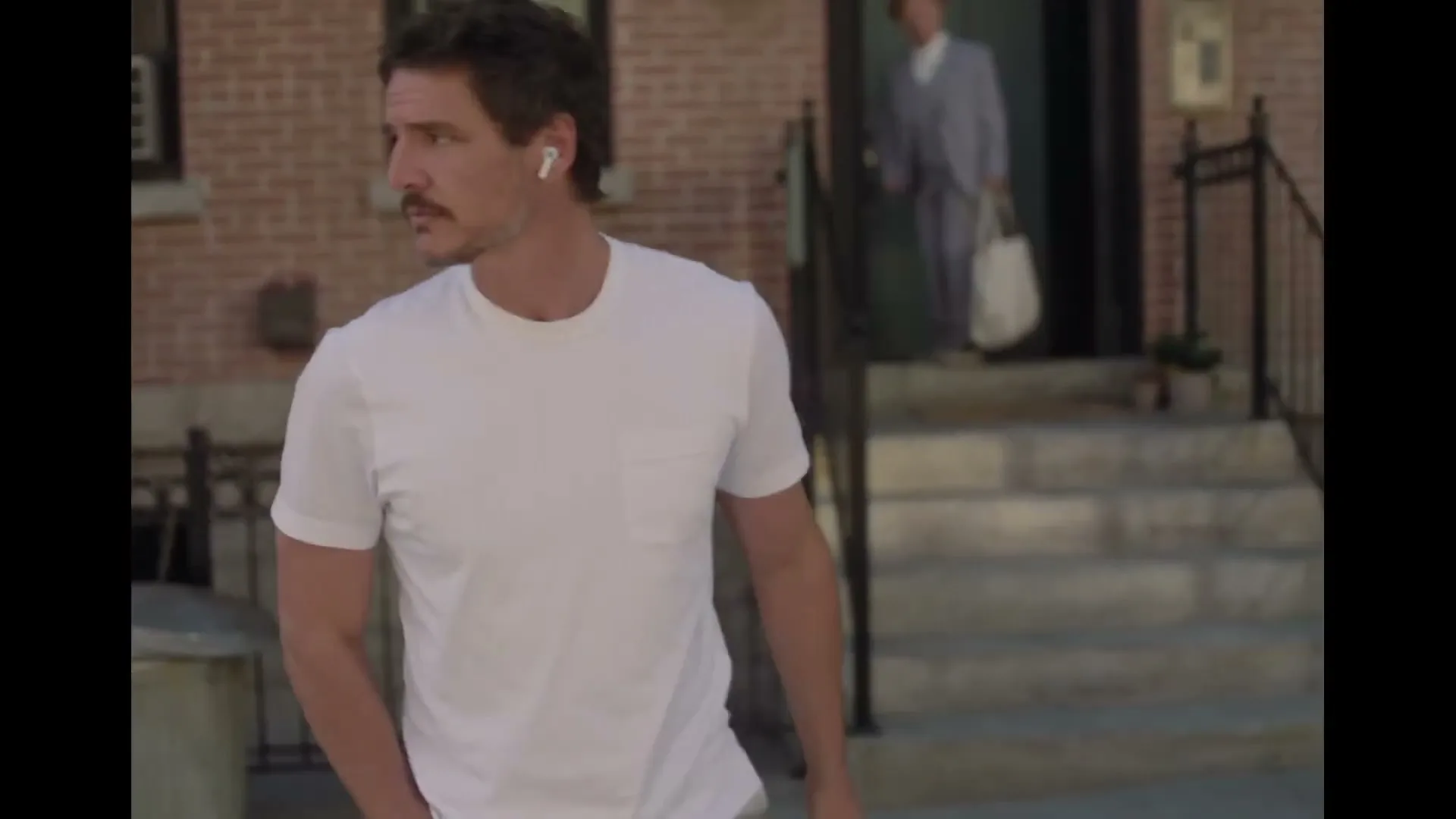 A man with a mustache and white t-shirt walks down a city street, looking around and adjusting his wireless earbuds.