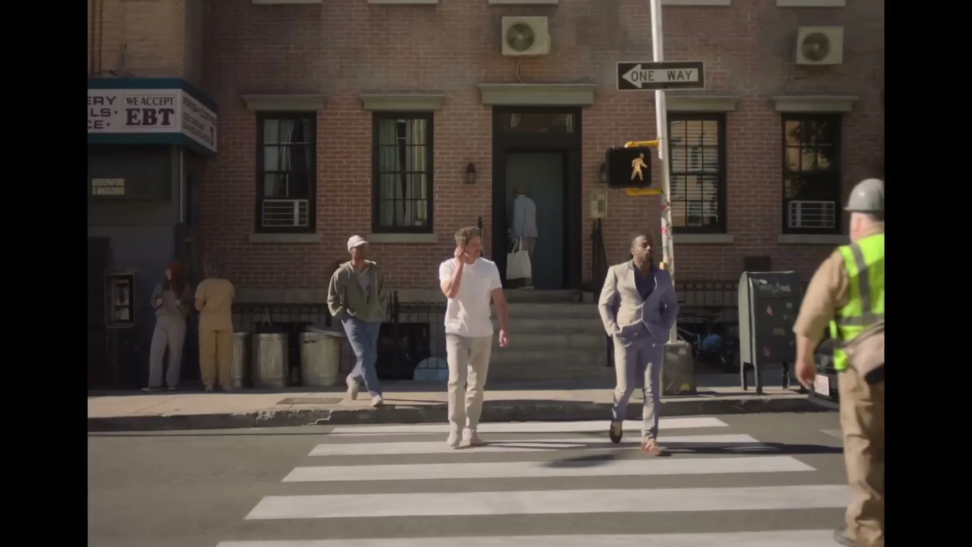 A diverse group of people cross a busy city street on a sunny day, with a man in a suit and another in casual wear walking towards the camera.