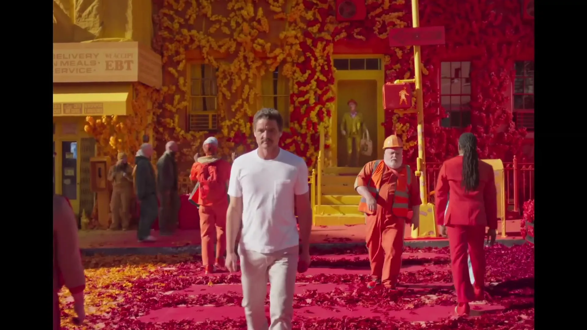 A man in a white t-shirt and light pants walks down a street covered in red petals, with buildings covered in yellow and red petals in the background.