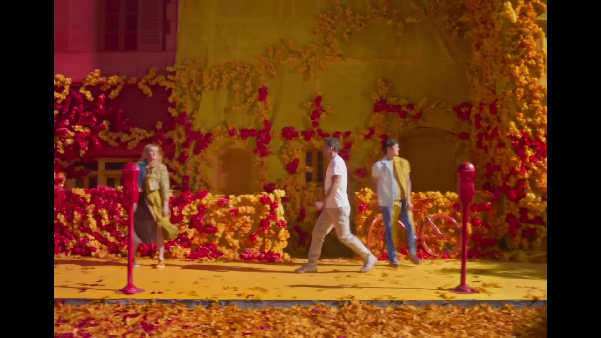 A diverse group of people walk past a building covered in yellow and red flowers, with a red bicycle parked on the sidewalk.