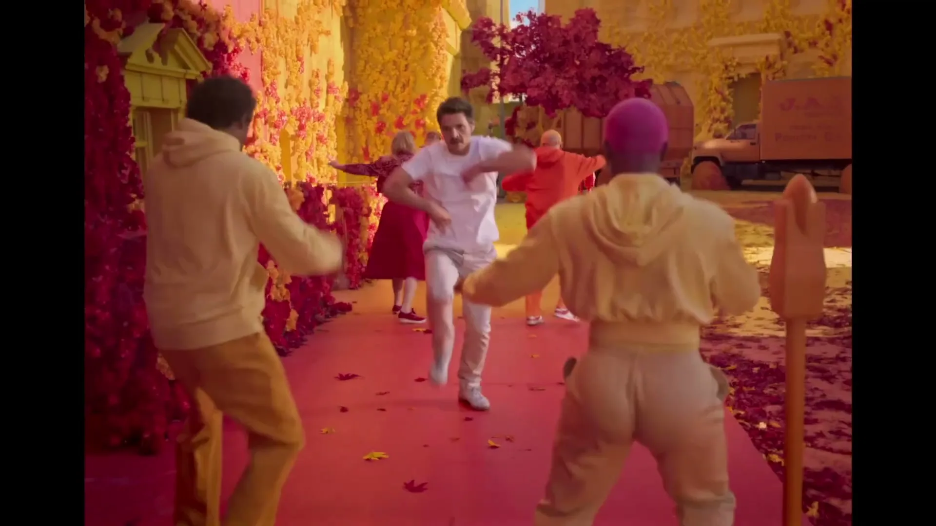 A man in a white t-shirt and light pants walks and dances down a red path, surrounded by people in colorful clothes and a vibrant, surreal landscape of red and yellow foliage.