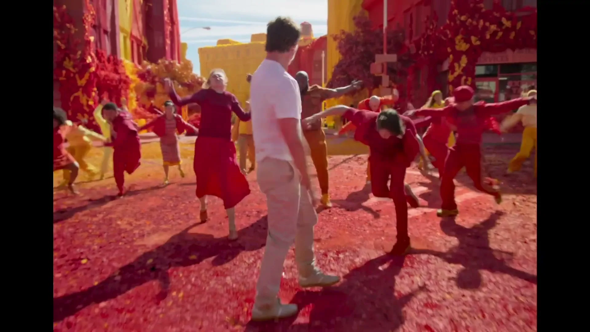 A man in a white t-shirt and khaki pants dances in the middle of a street, surrounded by a diverse group of people in red and yellow clothing, in a vibrant, color-saturated city.