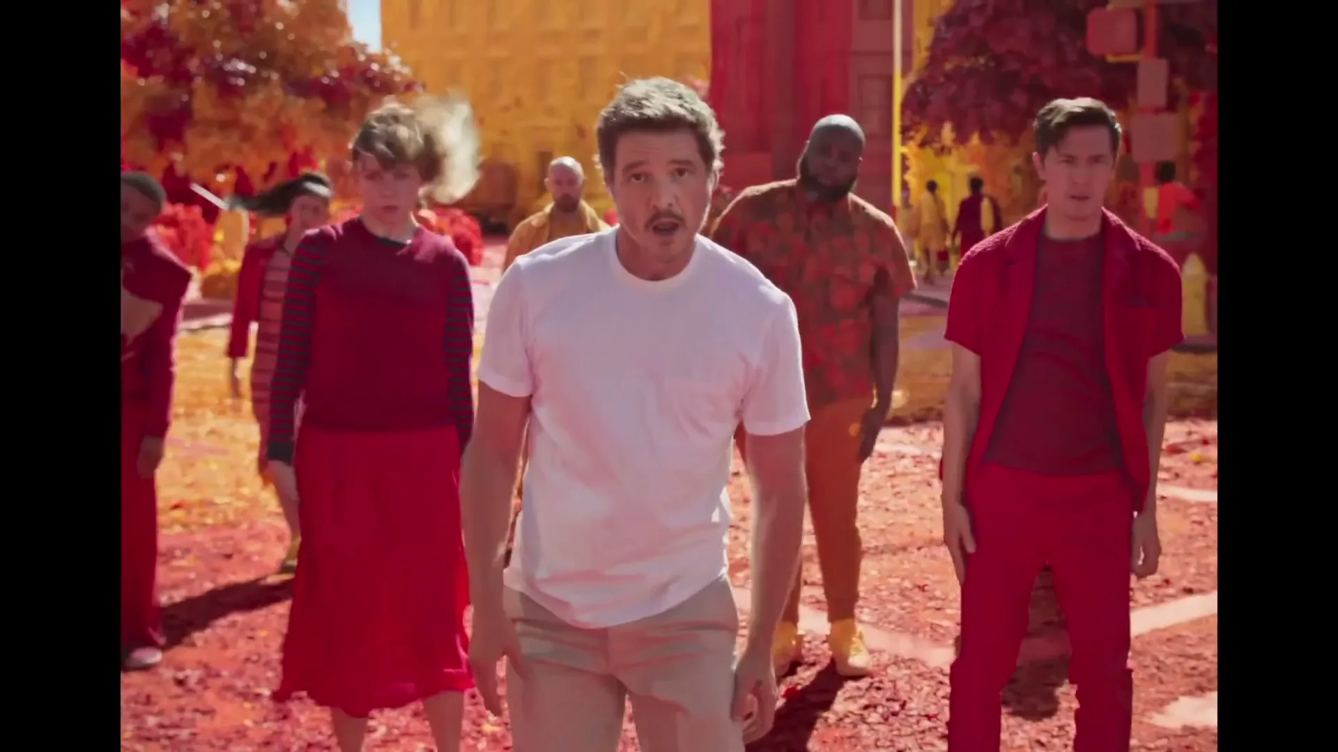 A man in a white t-shirt and beige pants dances with a group of people in a surreal, colorful outdoor setting.