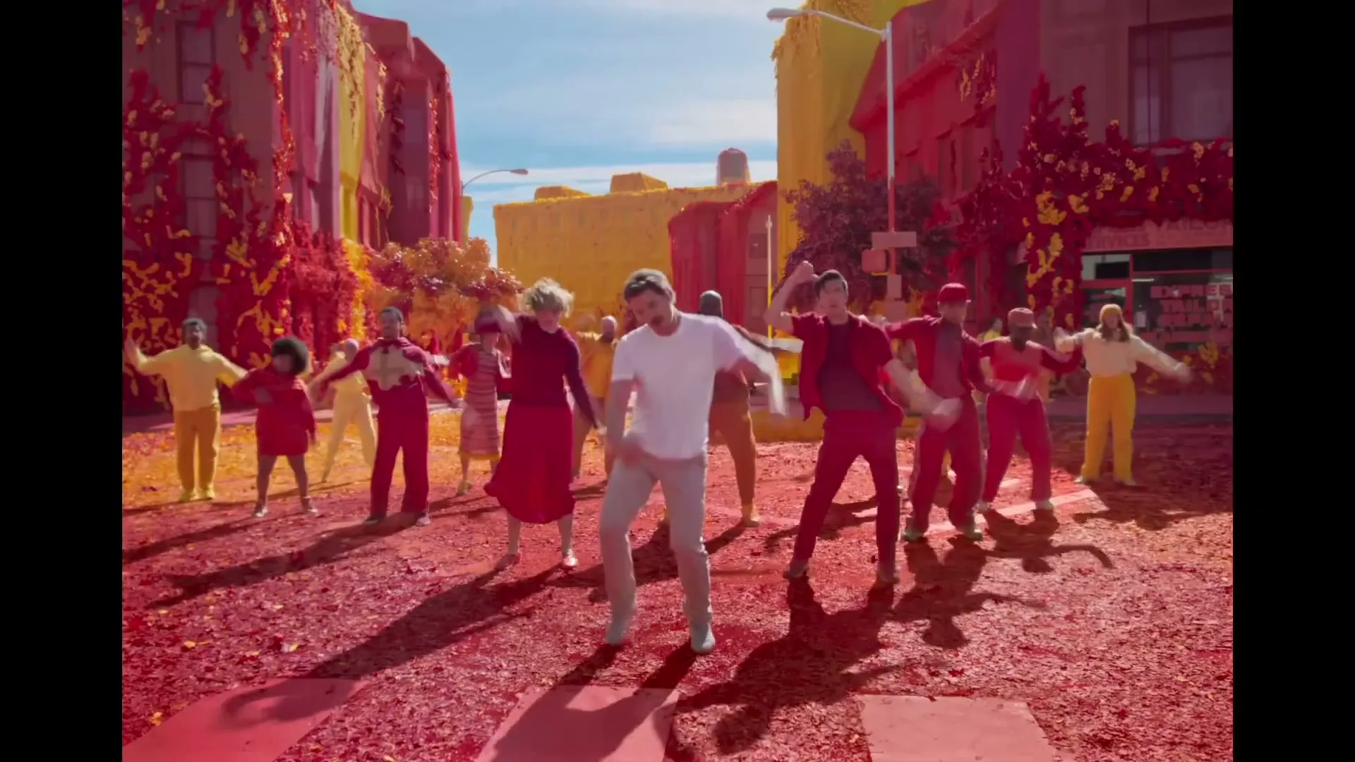A diverse group of people in colorful outfits dance in a street with buildings covered in red and yellow foliage, under a bright blue sky.
