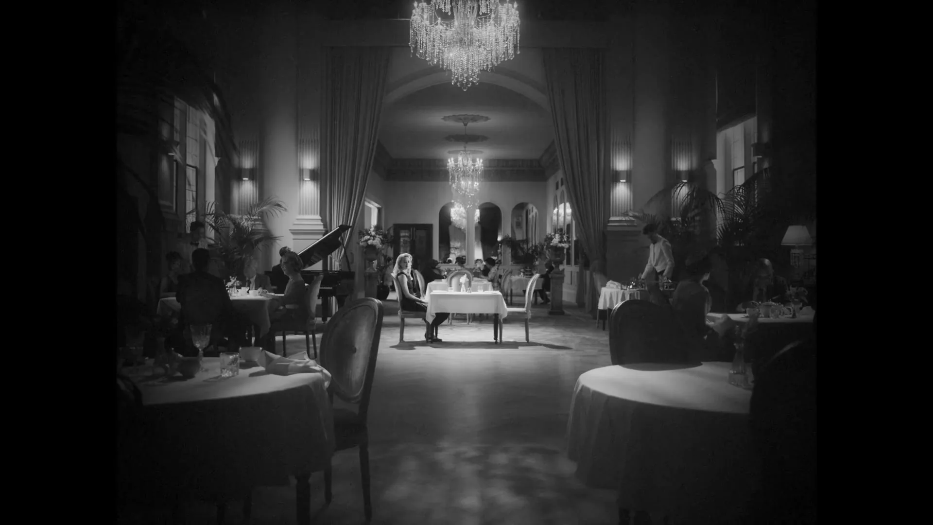 A woman with blonde hair sits alone at a table in a dimly lit, elegant restaurant, looking around, in black and white.