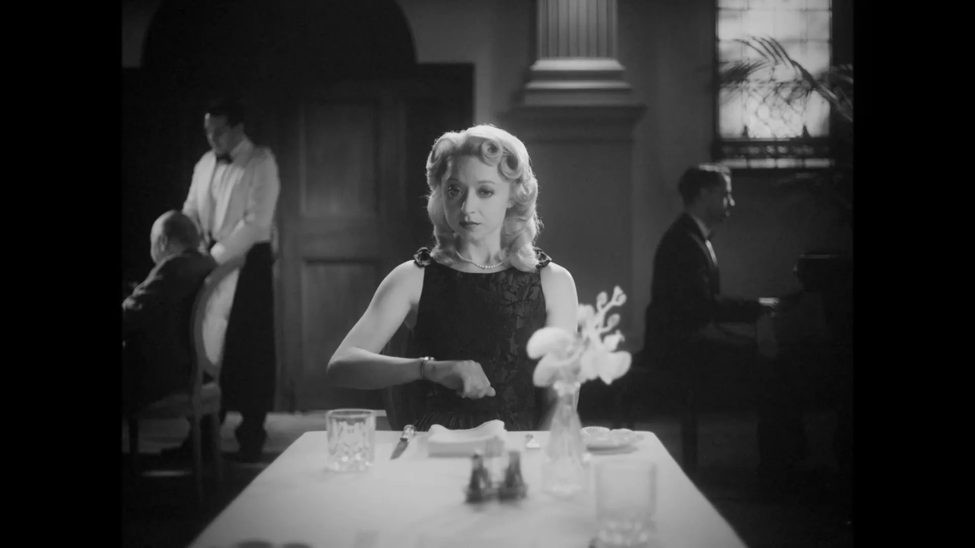 A woman with blonde hair in a black dress sits at a table in a restaurant, looking at her watch.