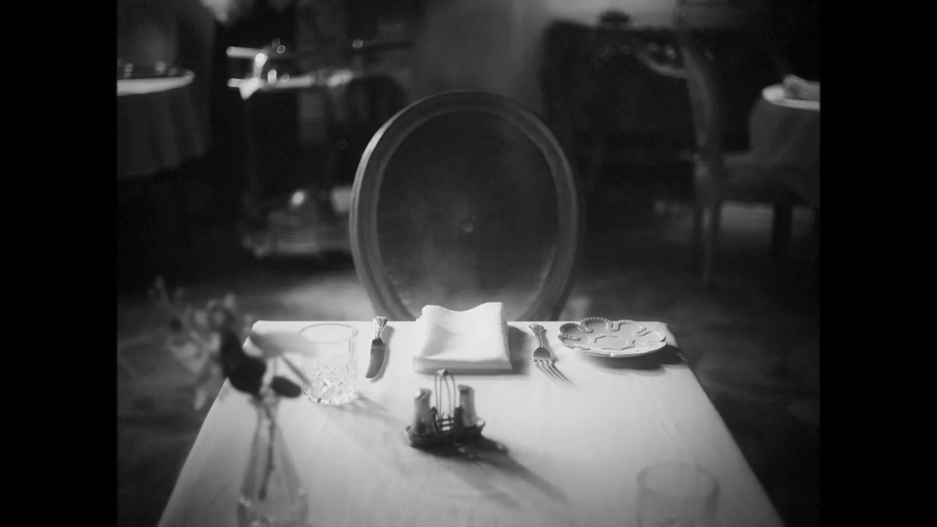 A black and white shot of a set table in a restaurant, with a chair pulled out, waiting for a diner.