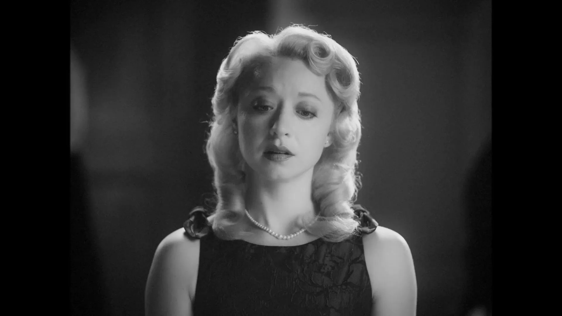 A blonde woman in a black dress and pearl necklace looks directly at the camera, then slowly lowers her gaze in a black and white shot.