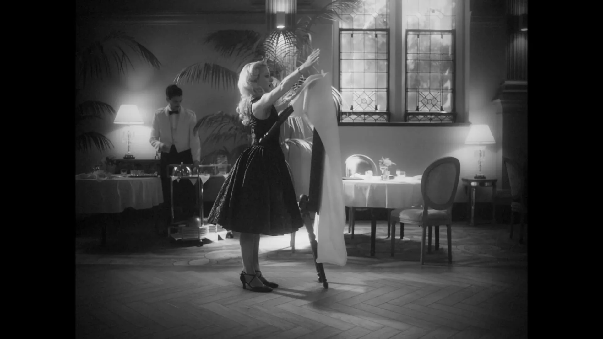 A woman in a black dress dances with a table and chair in a fancy restaurant, in black and white.