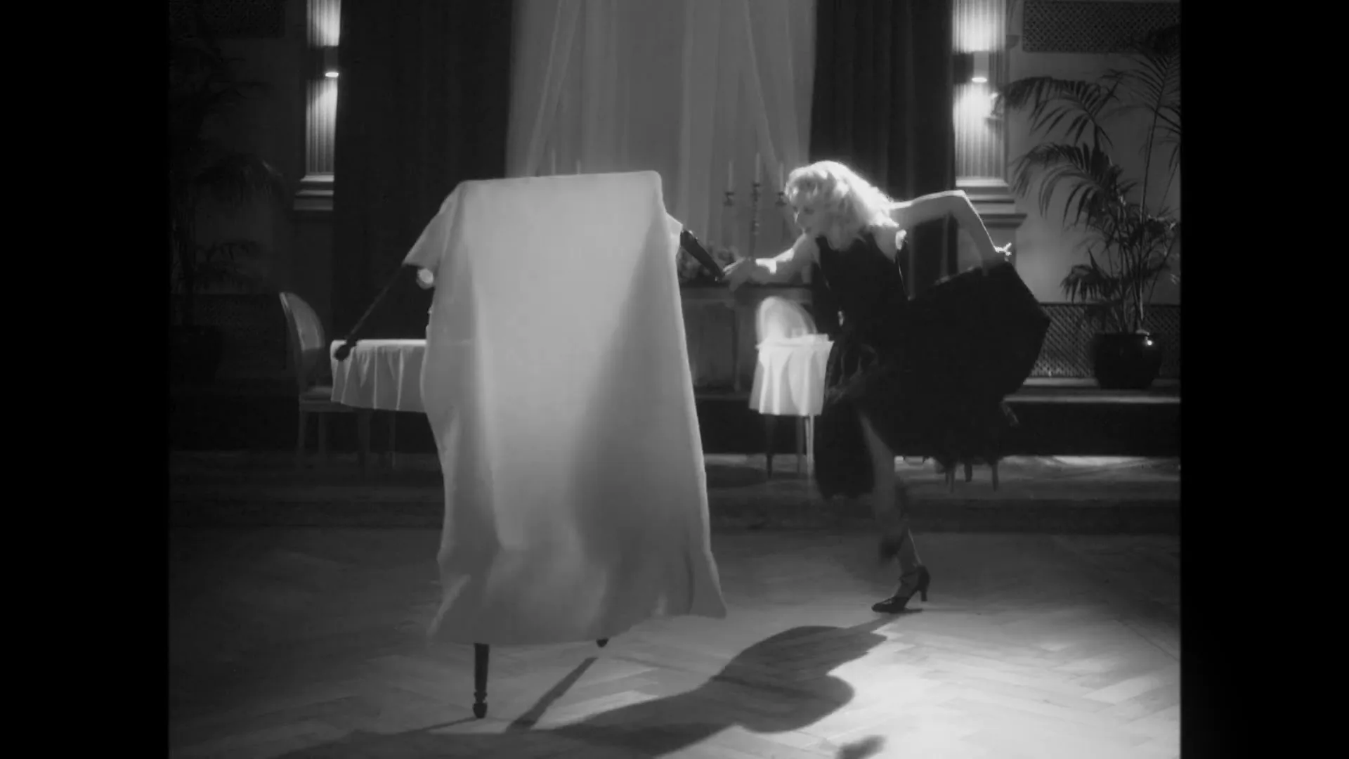 A blonde woman in a black dress dances with a chair in a fancy restaurant, in black and white.