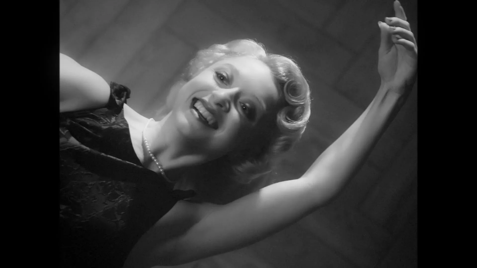A glamorous woman with blonde hair and a pearl necklace smiles and reaches up, looking down at the camera in a black and white shot.