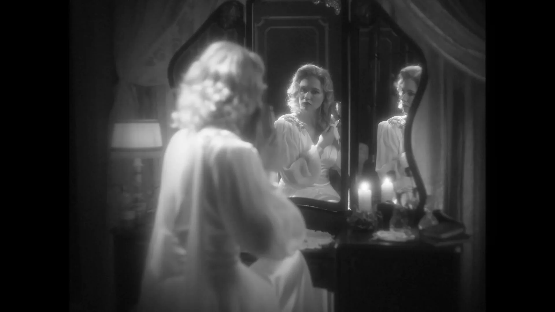 A woman with blonde hair in a white robe sits at a vanity table, looking at her reflection in a mirror in a dimly lit, opulent room.