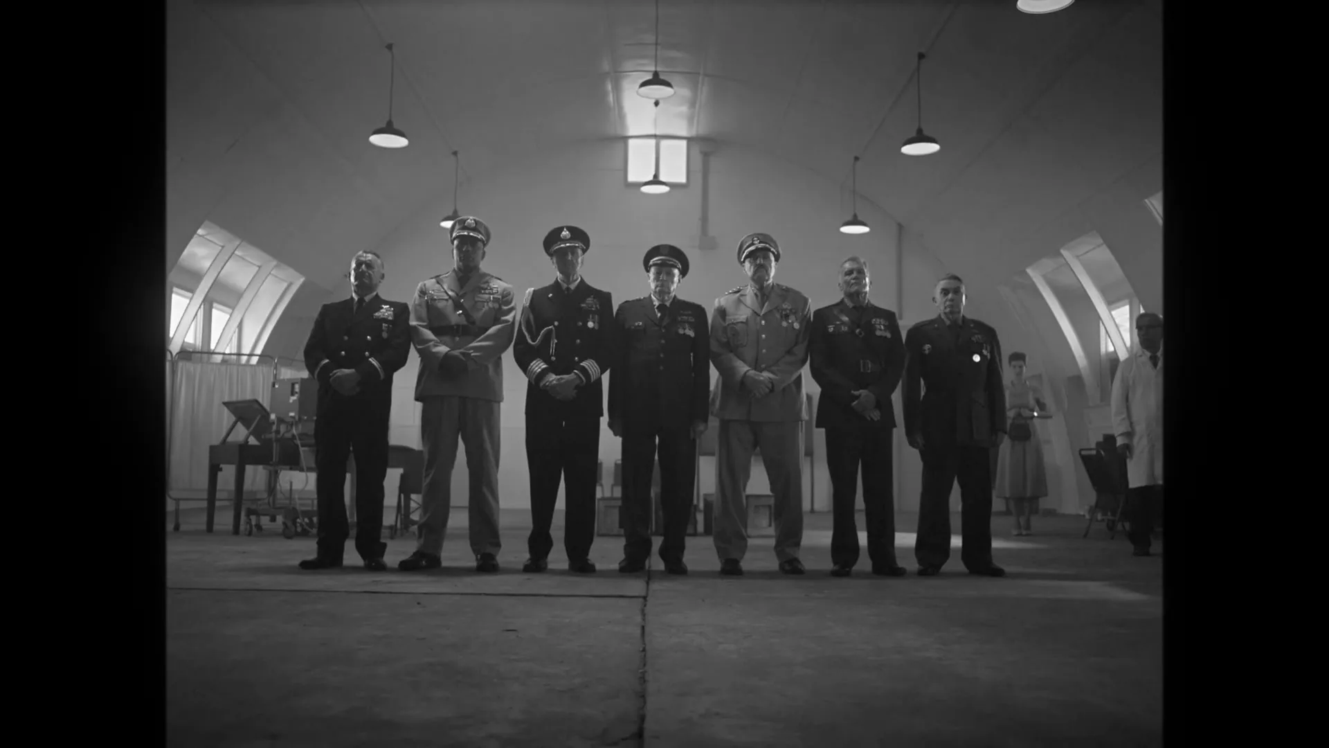 A young boy sits in a chair, facing away from the camera, as a group of military officers stand in a line in a large, empty room.