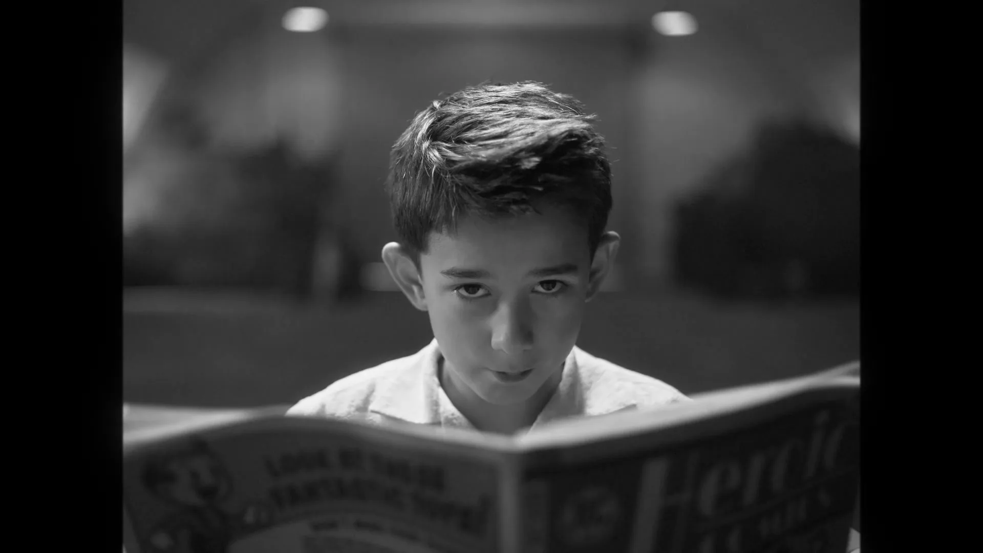 A young boy with short hair and a collared shirt reads a comic book, looking up briefly, in a dimly lit interior.